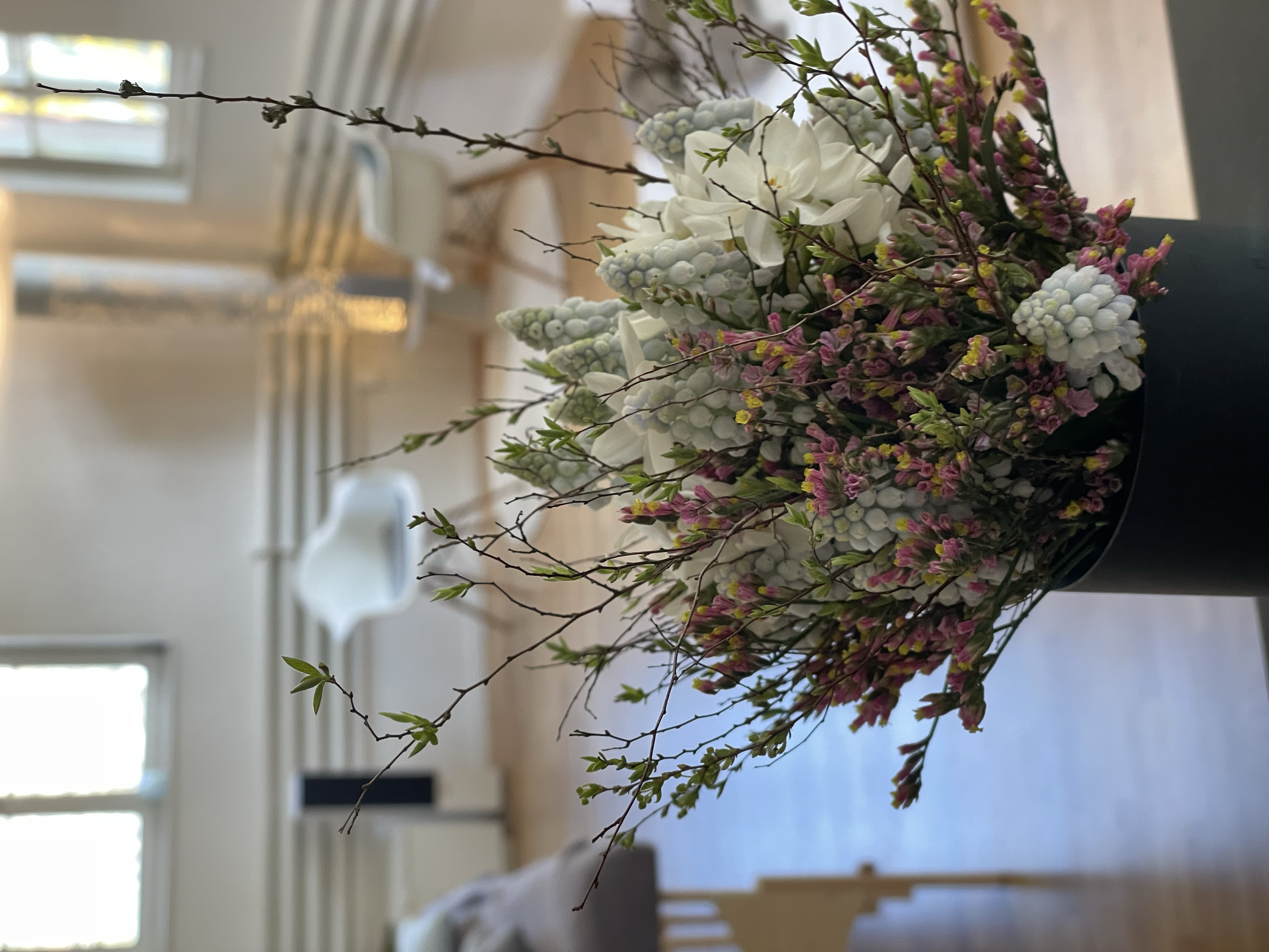 Blumenstrauß mit weißen, pinken und grünen Blumen in einem Raum mit natürlichen Licht, im Hintergrund ein Fenster und Stühle.