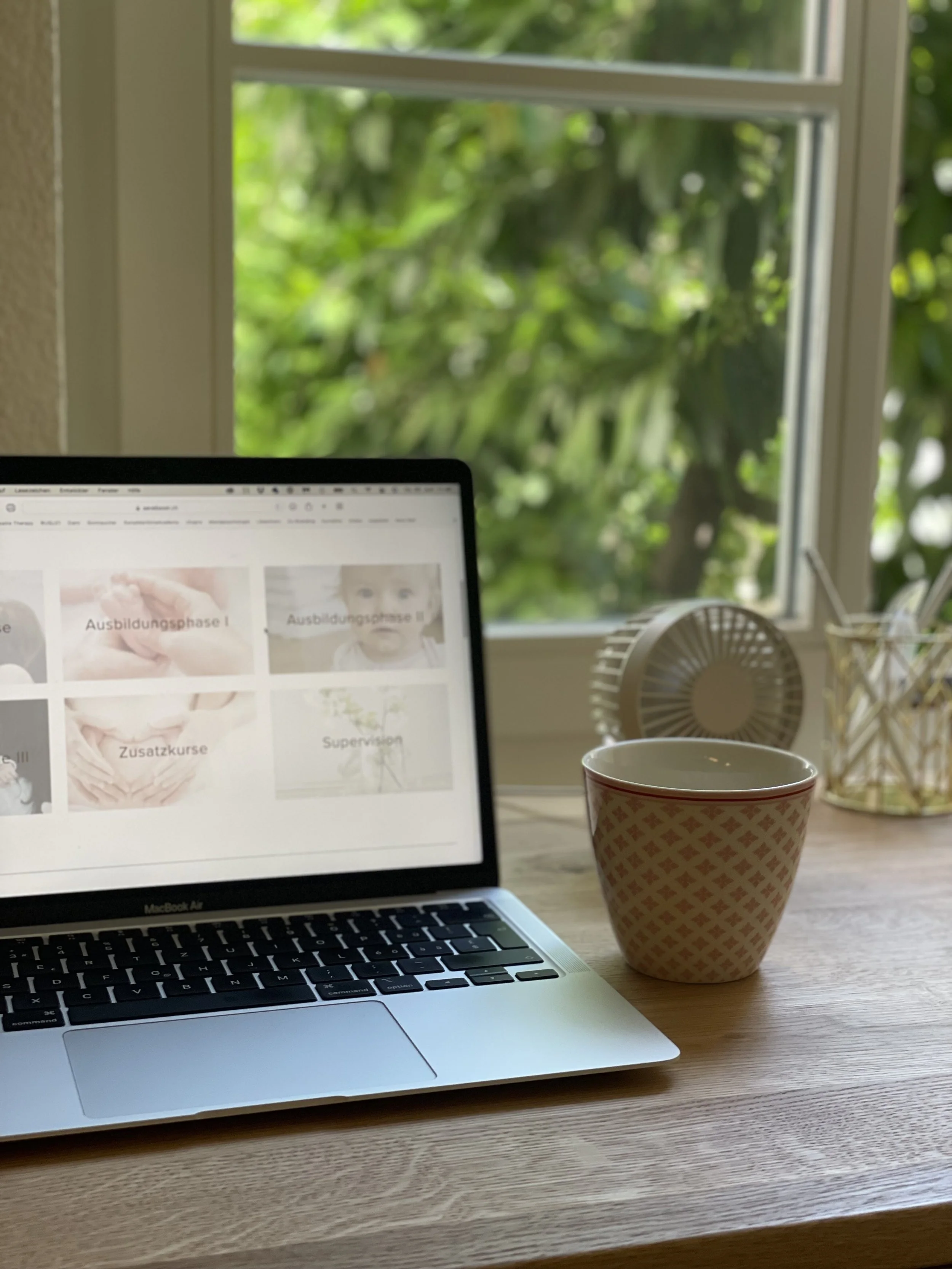 Der Blick auf einen Laptop auf einem Holzschreibtisch mit Kaffeetasse, ein Ventilator und Fenster mit grünen Bäumen im Hintergrund.