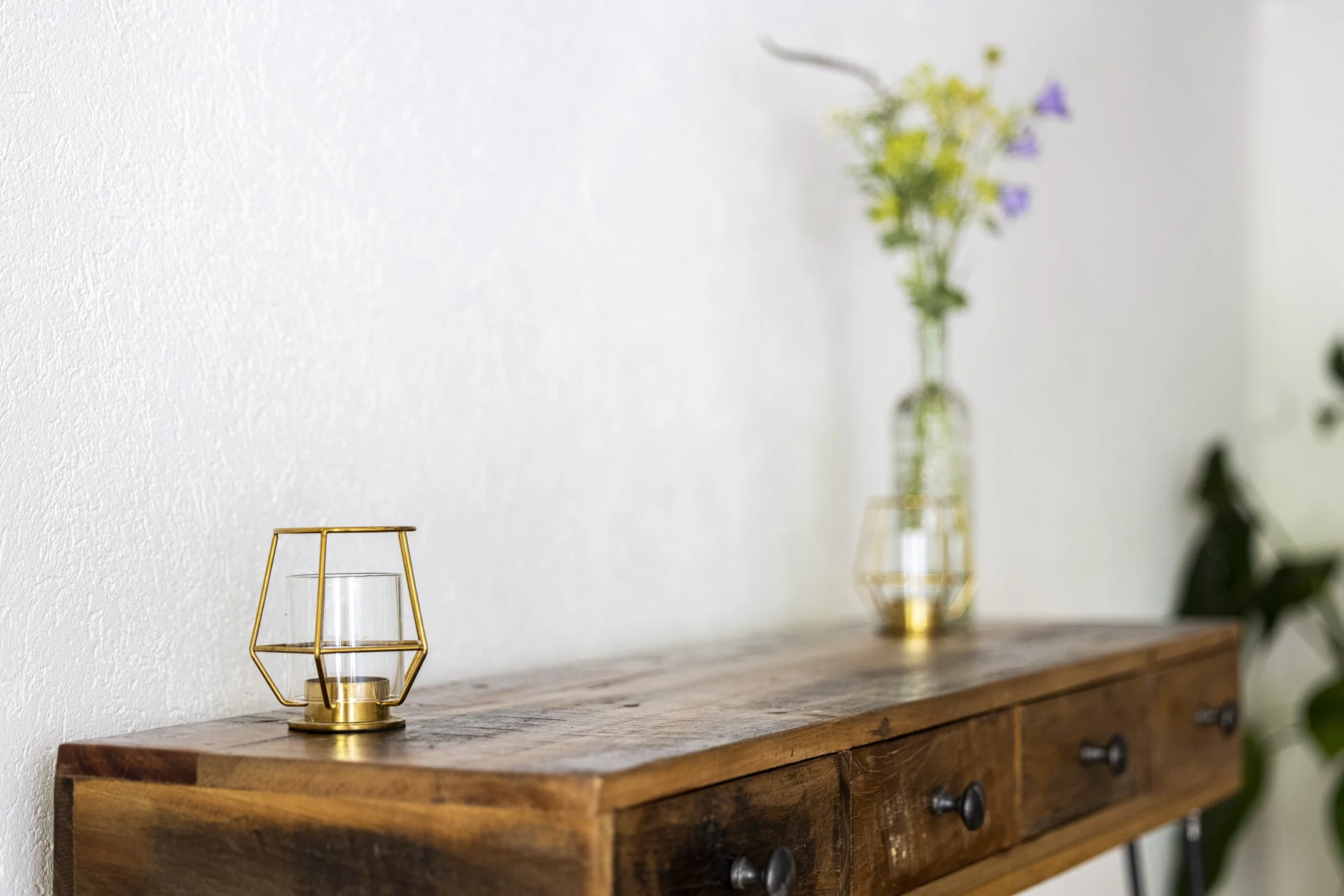 Holztisch mit einer goldenen Lampe und einer Vase mit Blumen vor weißer Wand.