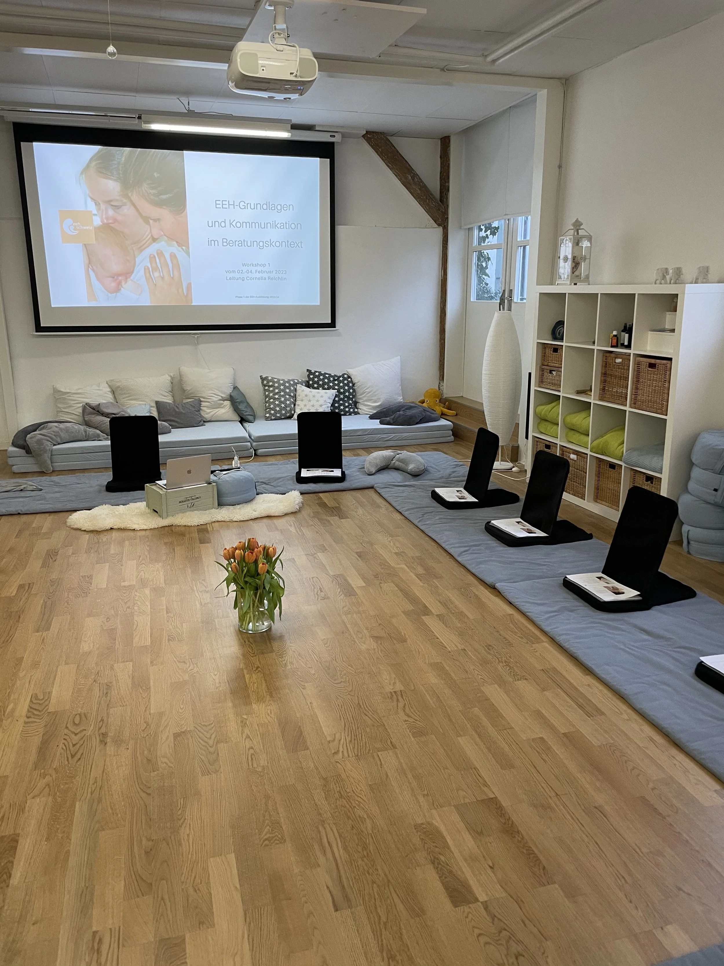 Ein Raum mit einem Projektor, einer großen Leinwand mit einer Präsentation, mehreren Sitzkissen auf dem Boden, einem Tisch mit einem Laptop, und einem Regal mit gefalteten Decken und anderen Gegenständen.