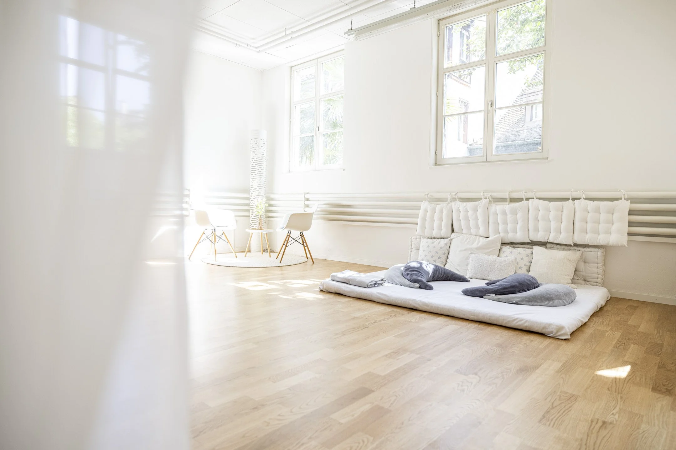 Ein helles, modernes Wohnzimmer mit großen Fenstern, weißen Wänden und Holzboden. Es gibt einen weißen Sofaboden mit Kissen und eine kleine Ecke mit Stühlen und einem Teppich.