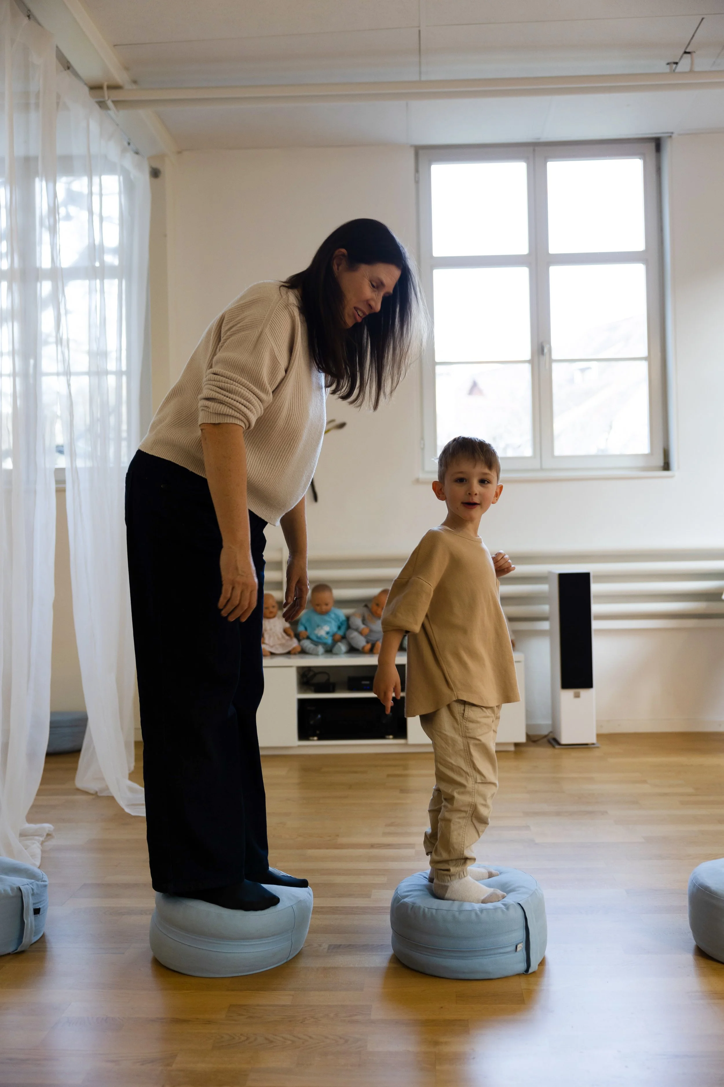 Eine Frau und ein Junge stehen auf runden Kissen auf einem Holzboden. Sie scheinen an einer Balance-Aktivität teilzunehmen, während im Hintergrund Babys auf einer Couch sitzen. Das Zimmer ist hell mit großen Fenstern.