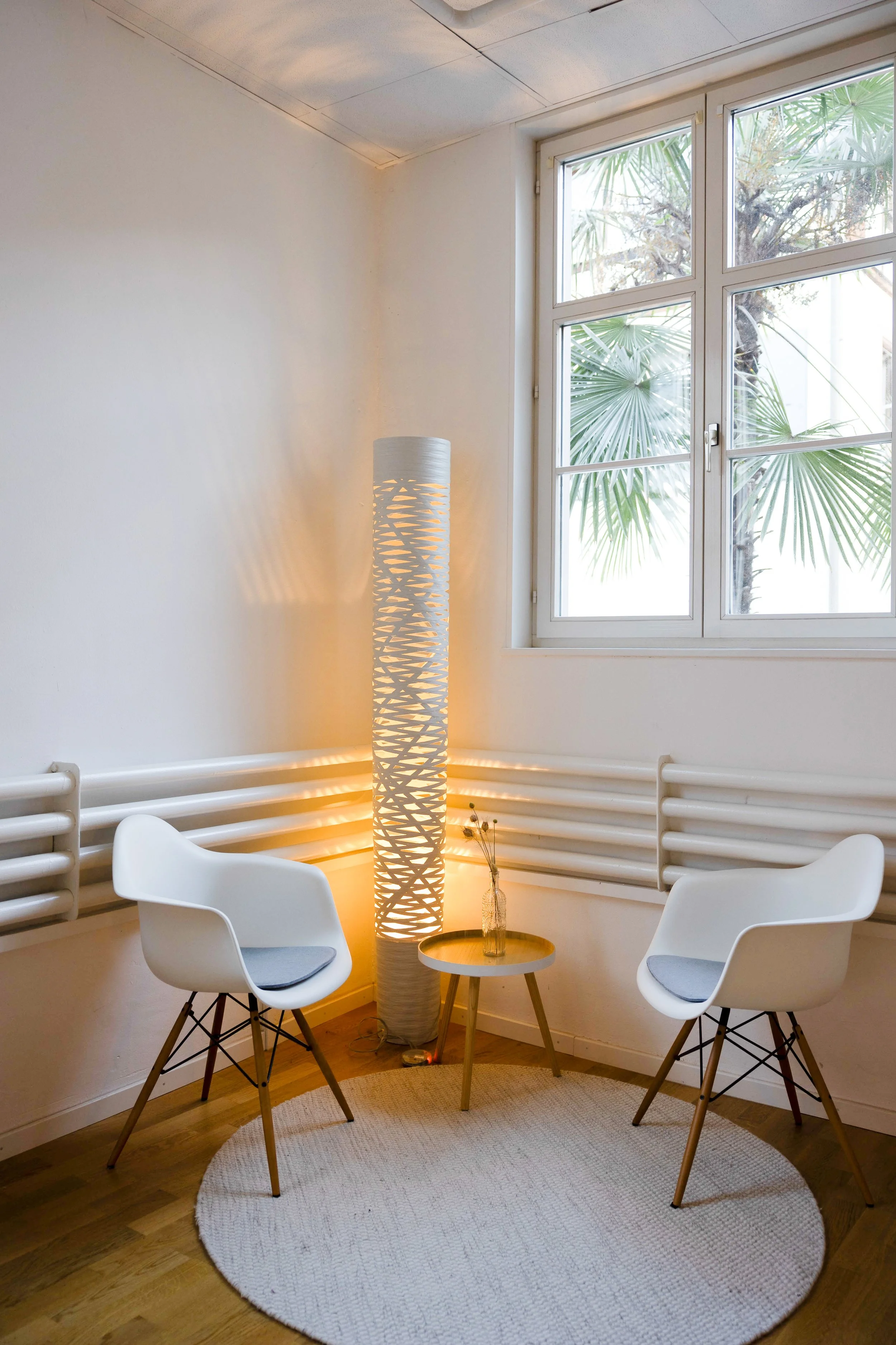 Ein gemütlicher Sitzbereich mit zwei weißen modernen Stühlen, einem kleinen runden Beistelltisch mit einer Vase, einem hohen Stehlampe mit designierter Struktur, einem großen Fenster mit Blick auf Palmen, einem runden, hellen Teppich auf Holzboden un