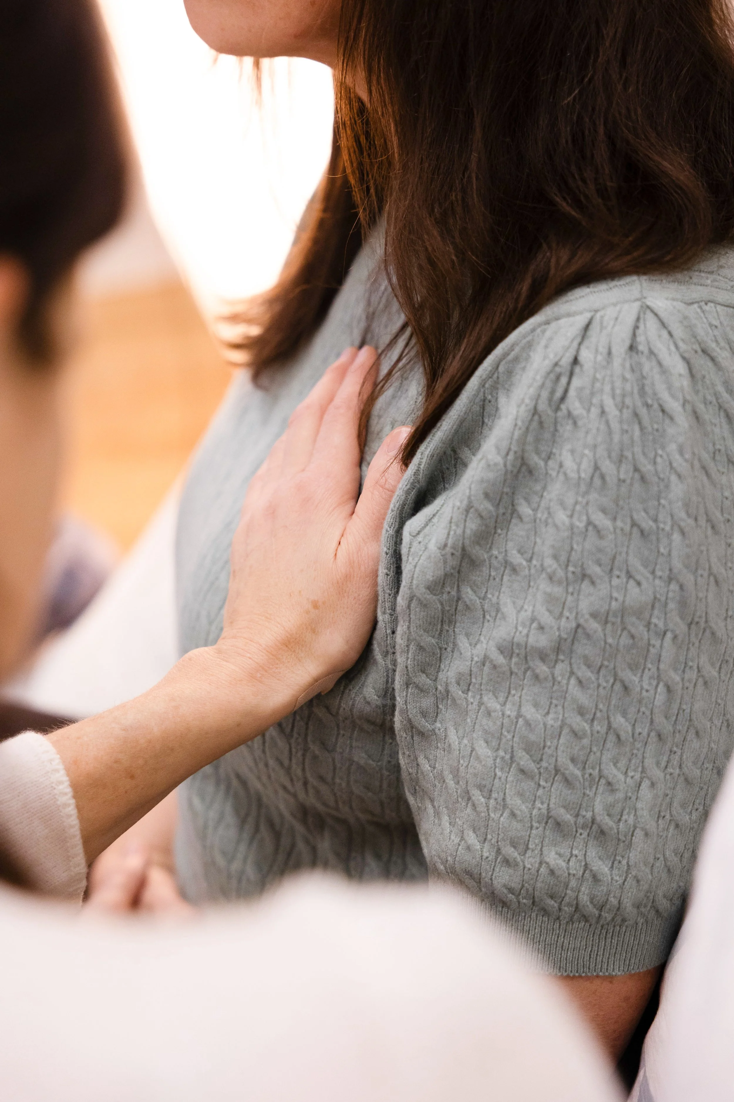 Eine Nahaufnahme zeigt eine Hand, die auf die Schulter einer Frau mit braunen Haaren und grauem Pullover gelegt ist.