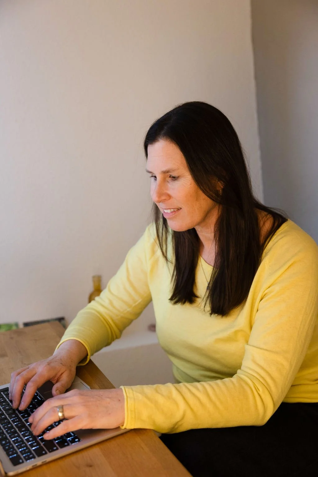 Eine Frau mit schwarzen Haaren, gelbem Pullover, arbeitet an einem Laptop auf einem Holztisch, im Hintergrund eine weiße Wand.