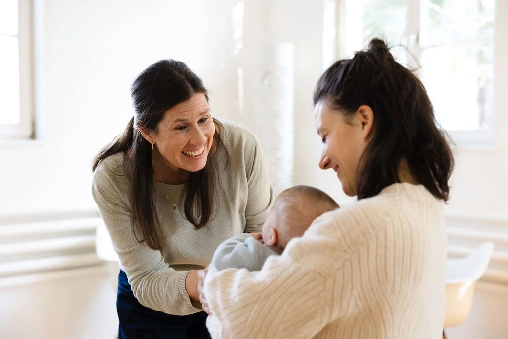 Zwei Frauen halten ein Baby, lachen und schauen sich liebevoll an.