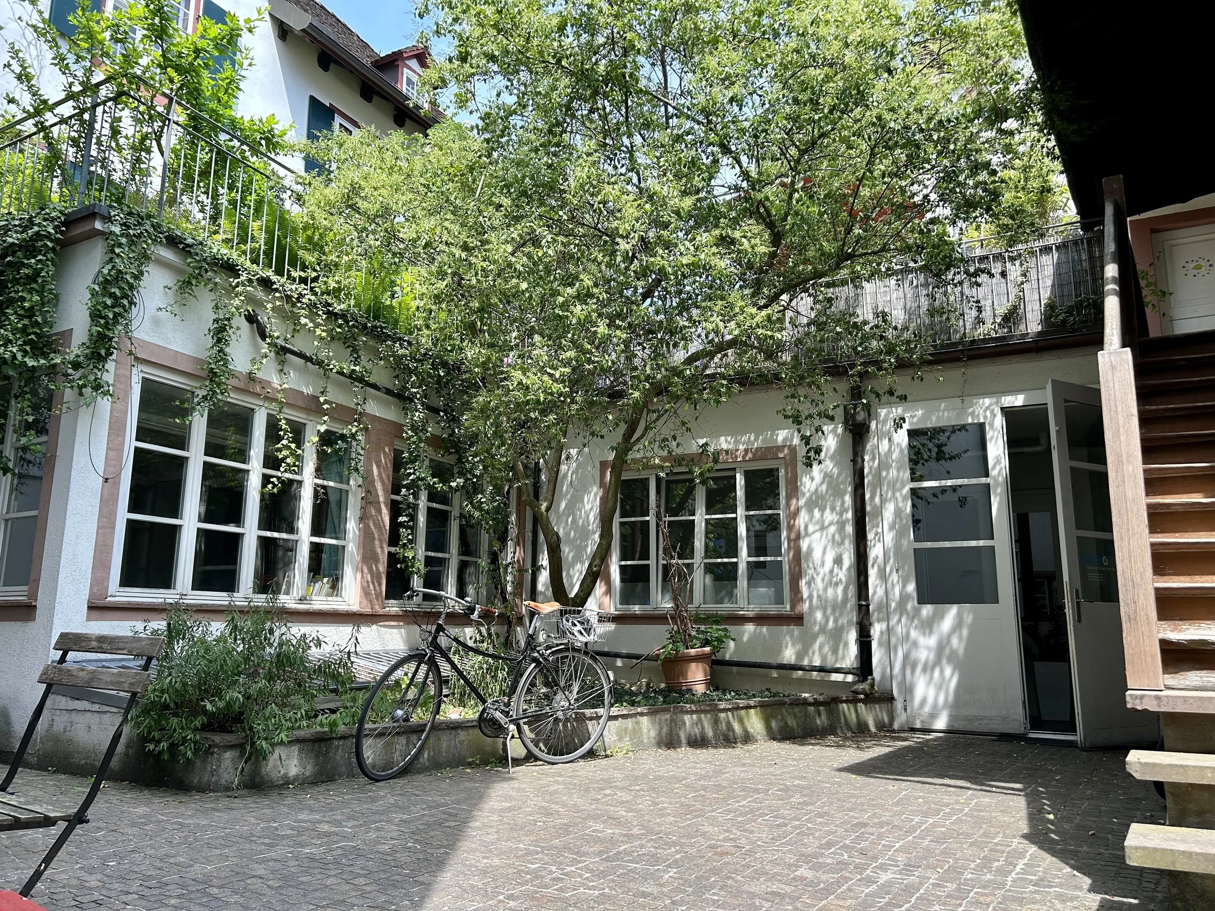 Ein Innenhof mit einem Baum in der Mitte, Fahrrädern, einer Bank und einem Steingarten im Vordergrund; im Hintergrund ein Haus mit weißen Wänden und mehreren Fenstern, umgeben von viel Grün.