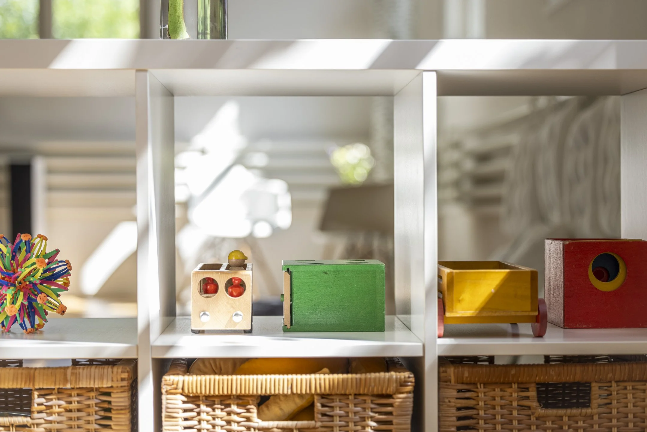 Ein weißes Regal mit bunten Holzspielzeugen, Bällen, Bildelementen in Körben und anderen Spielsachen in einem Kinderzimmer.