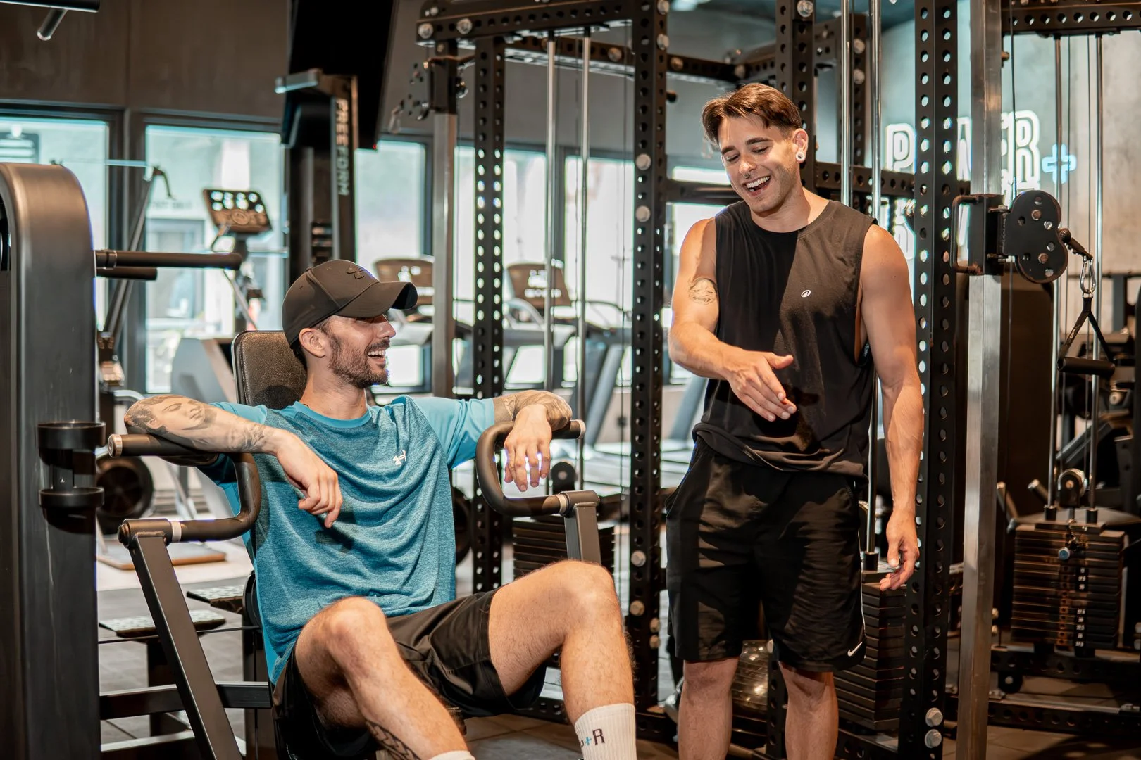 Two men in a gym, smiling and talking. One is sitting on a workout machine, wearing a blue shirt and black shorts, with tattoos and a black cap. The other stands beside him, wearing a black sleeveless shirt and shorts, gesturing with his hand.