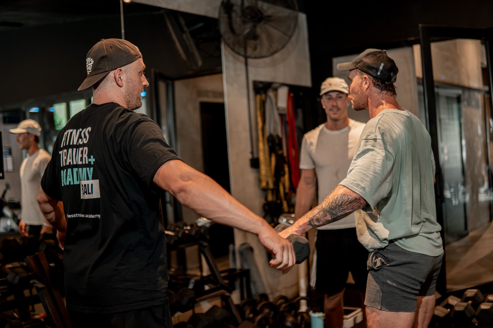 Four men in a gym. One man is holding a dumbbell and assisting another man with his workout. Two men are in the background, watching the interaction. The scene appears to be a fitness training session.