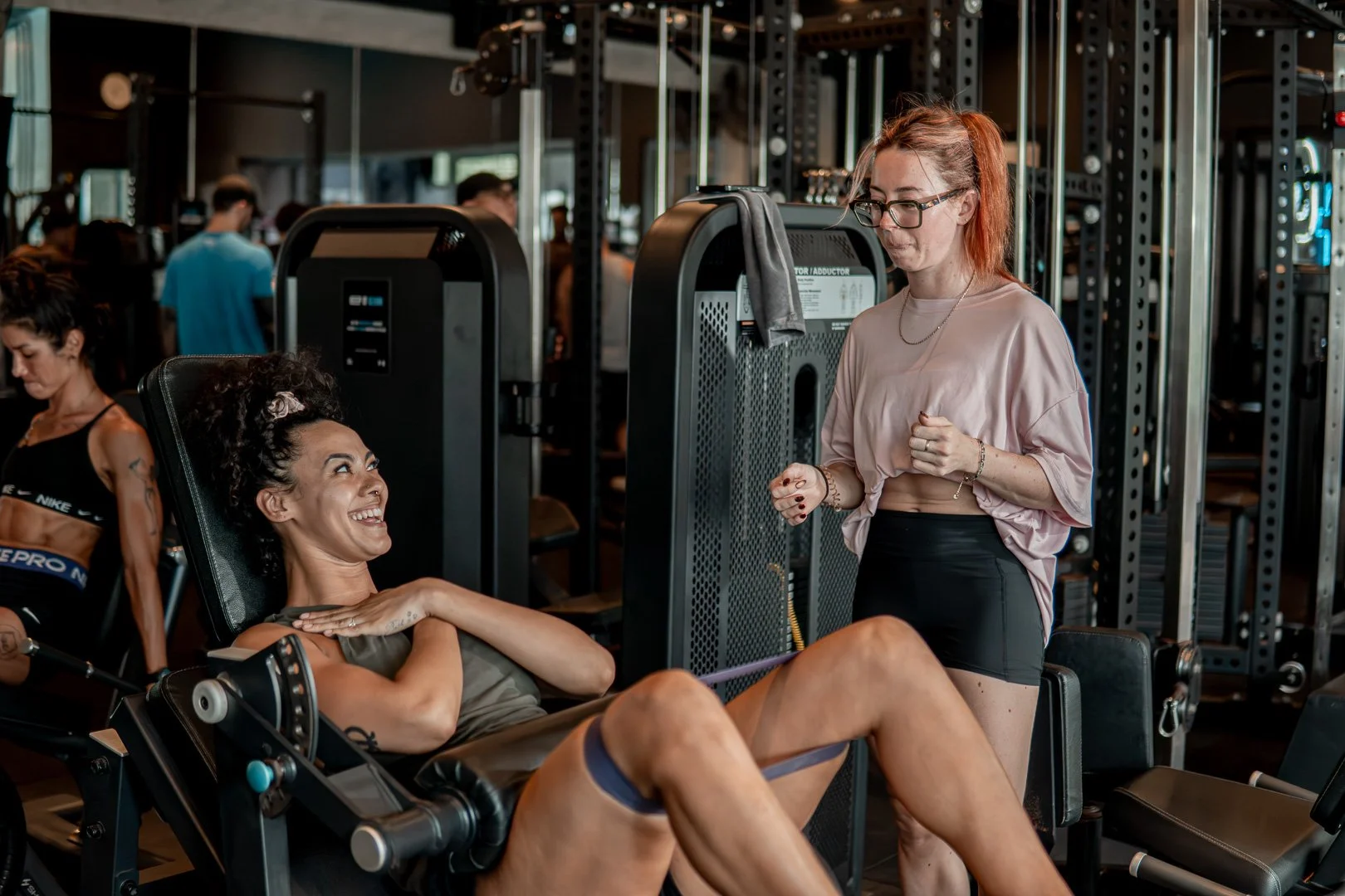 A woman with curly hair and athletic attire lying on a gym workout chair, smiling and looking at a woman with red hair and glasses standing beside her, in a gym environment with other people exercising in the background.