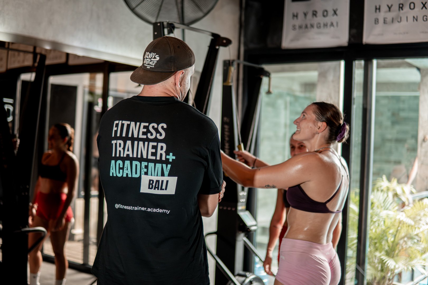 A fitness trainer in a black shirt talks to a woman in workout clothes at a gym, with others exercising in the background.