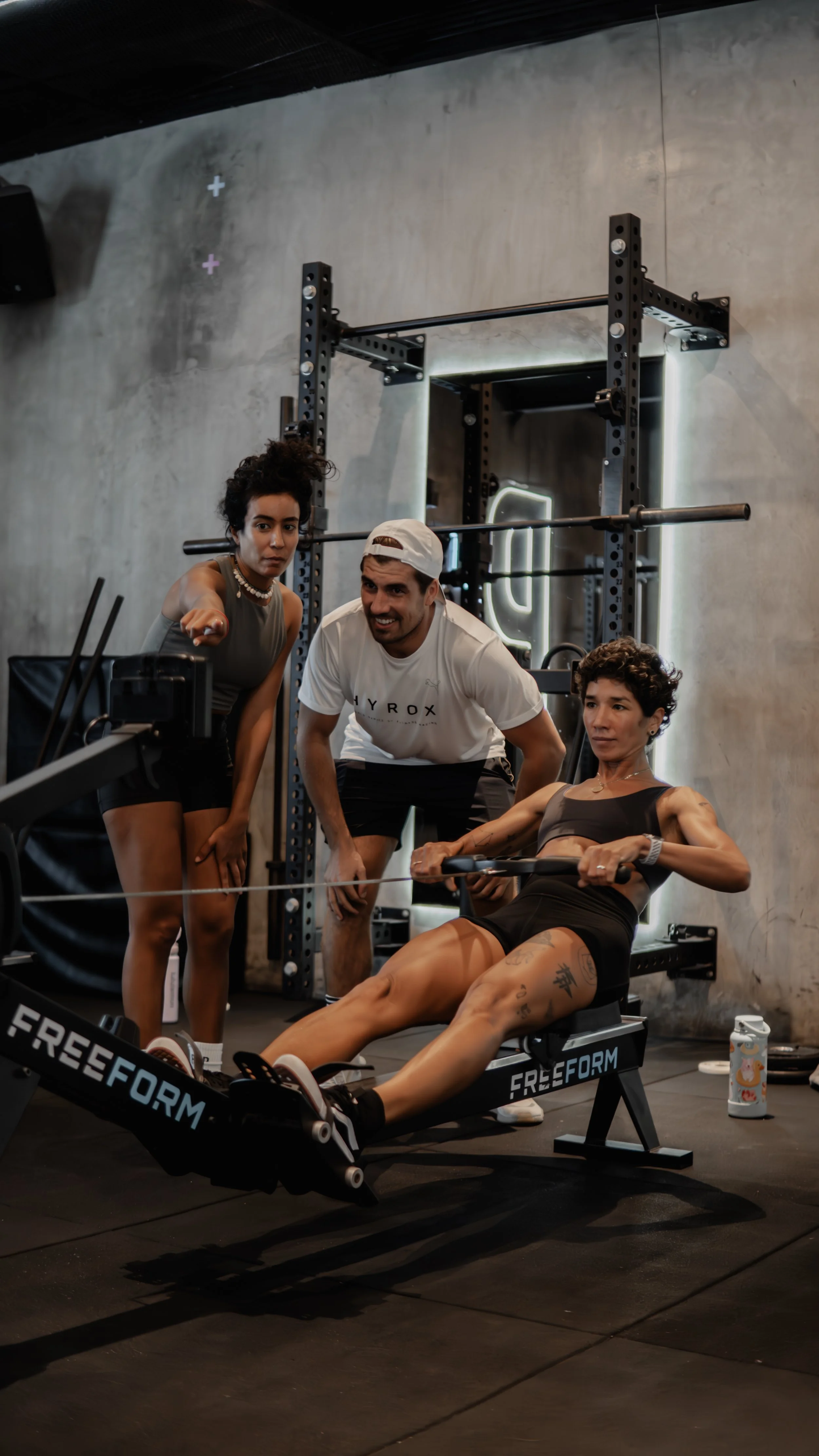 A woman is exercising on a rowing machine while two coaches or trainers observe and give instructions at a gym.