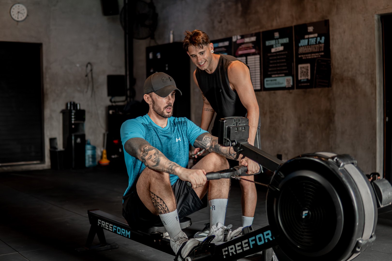 A man using a rowing machine while a trainer provides guidance in a gym.