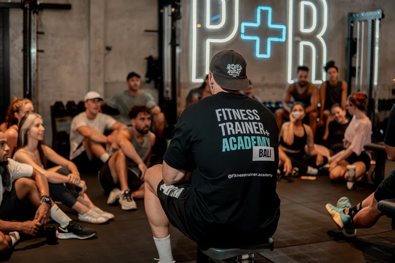 Fitness trainer leading a session with a group of people in a gym, sitting on the floor listening attentively, with a neon sign in the background reading 'P+R' and a cross symbol.