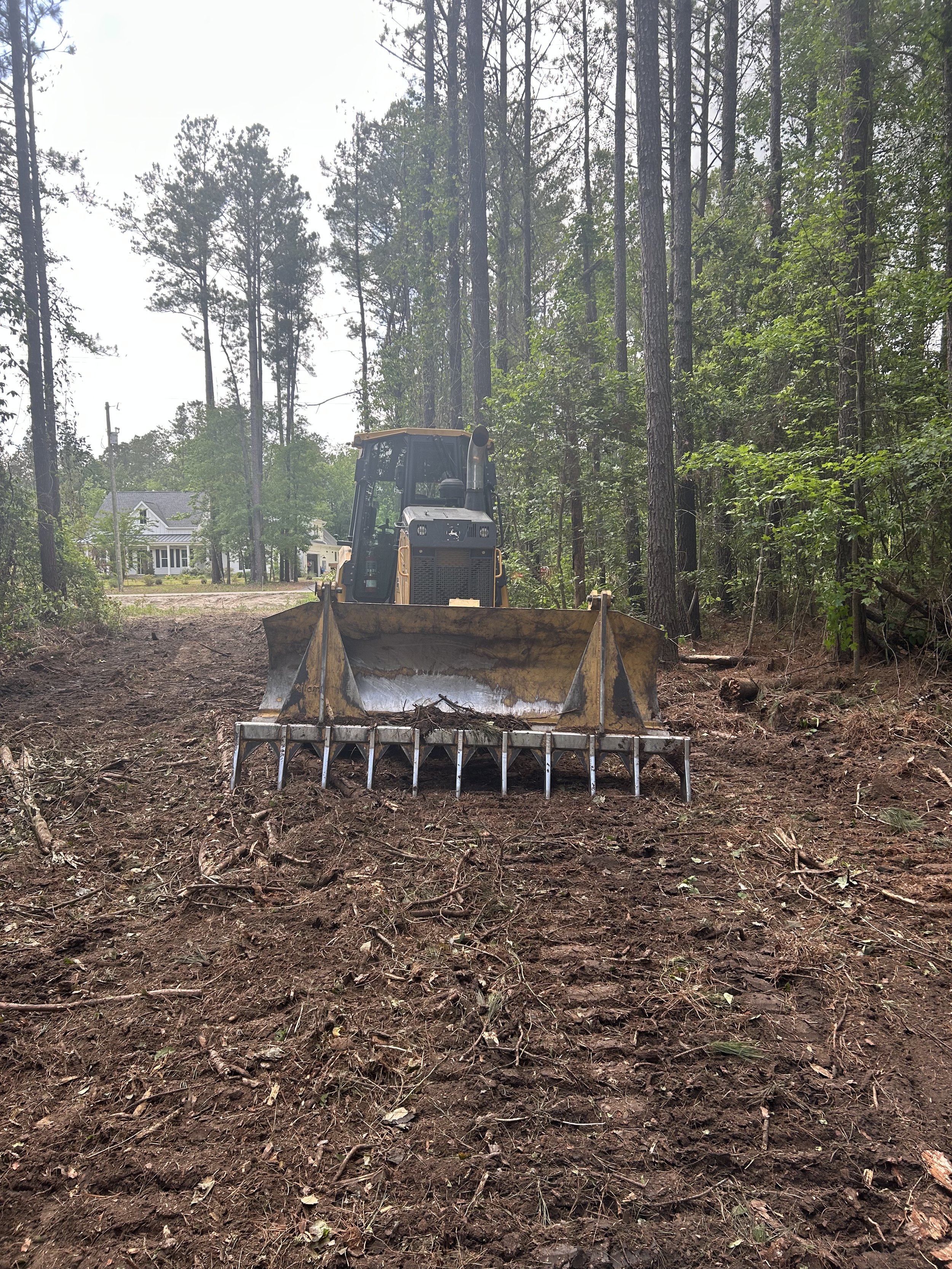 A bulldozer clearing a wooded lot with trees and a house visible in the background.