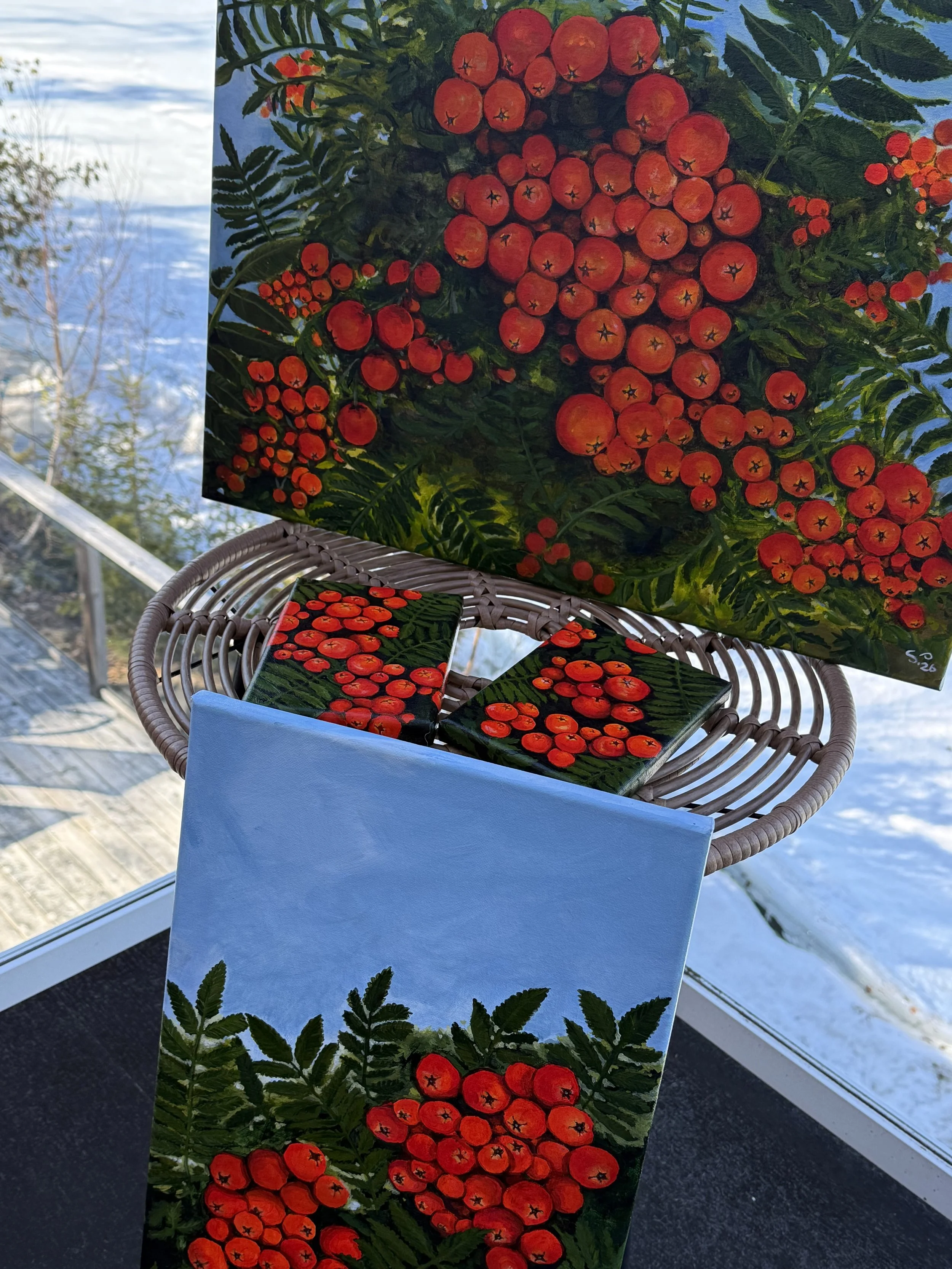 Artistic painting of red rowan berries, green leaves, and fern-like plant details, laid out on a surface with a smaller version of the same artwork, on a wicker table in sunlight.