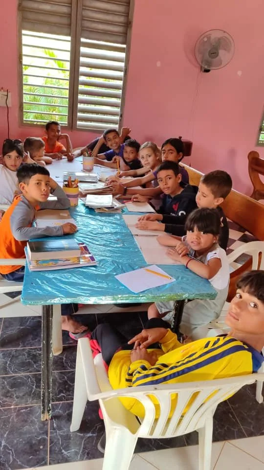 Group of children and a young woman seated around a table with school supplies, in a room with pink walls and large windows.