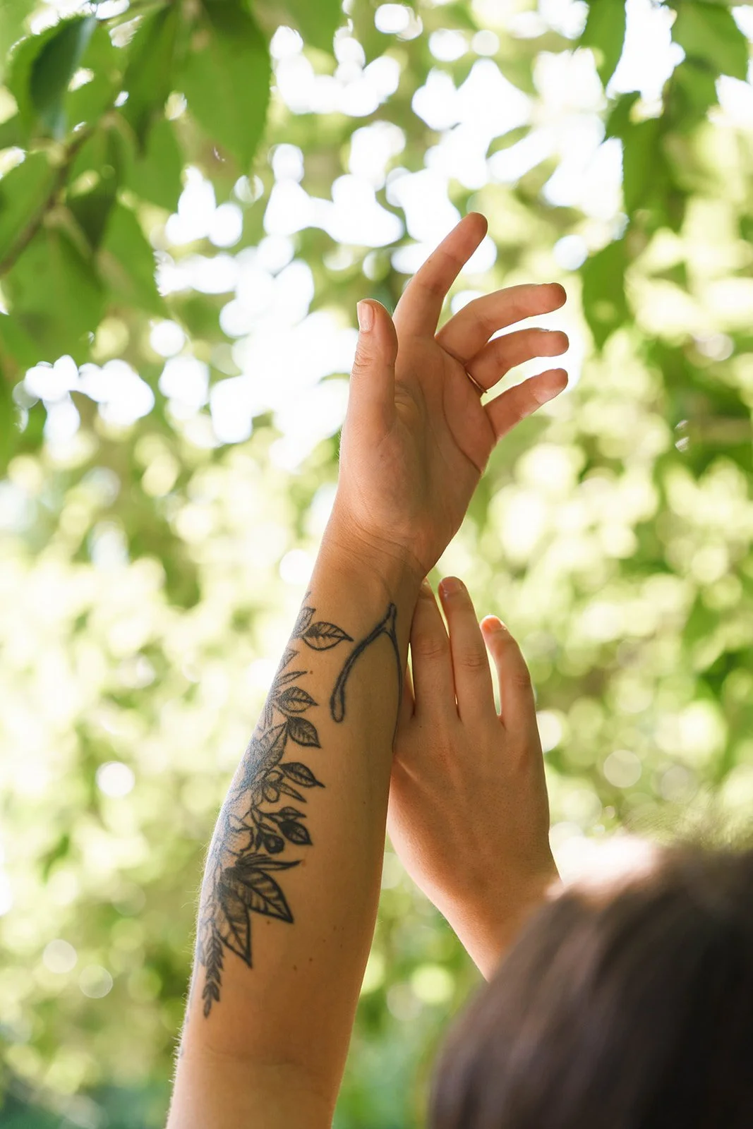 A person with a tattoo on their arm reaches upward with their fingers, surrounded by green leaves and dappled sunlight.