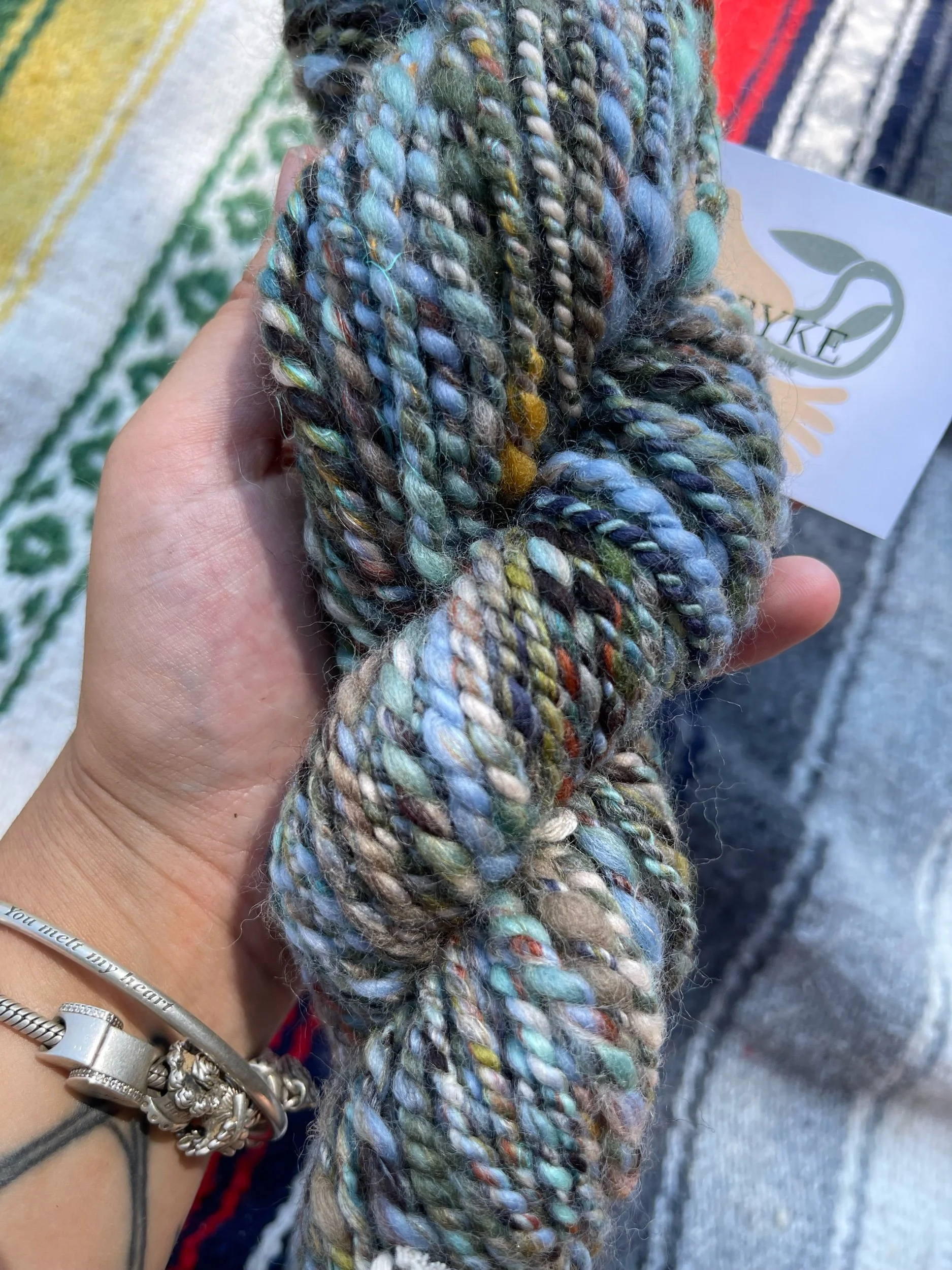 A hand holding a skein of multicolored, twisted yarn. The yarn is a mix of pastel and natural tones. There is a bracelet on the wrist with engravings and a charm. In the background, there are textile fabrics and a card with a leaf logo.
