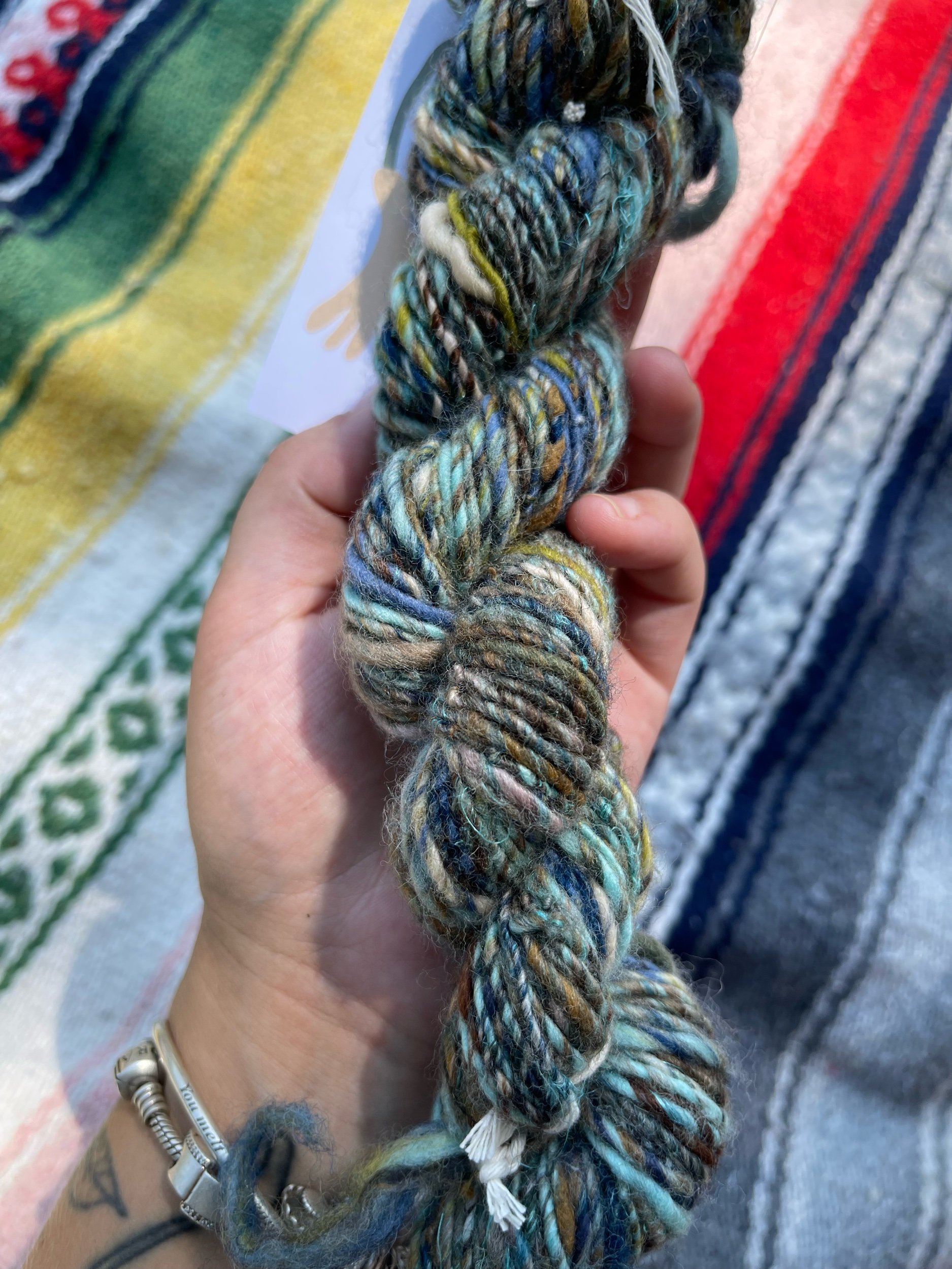 A hand holding a multicolored skein of yarn with a embroidered blanket in the background.