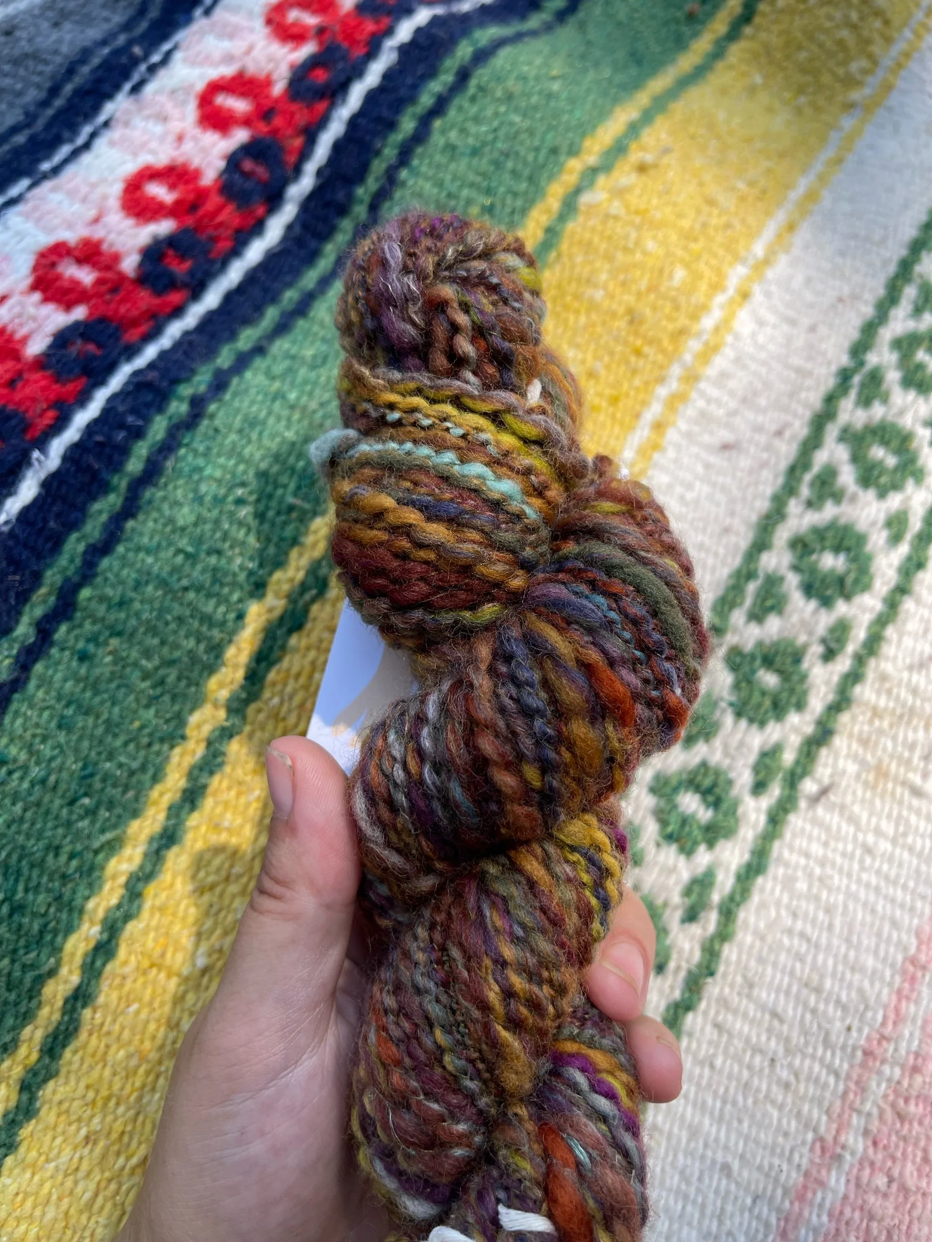 Close-up of a hand holding a multicolored skein of yarn over a colorful woven textile background.