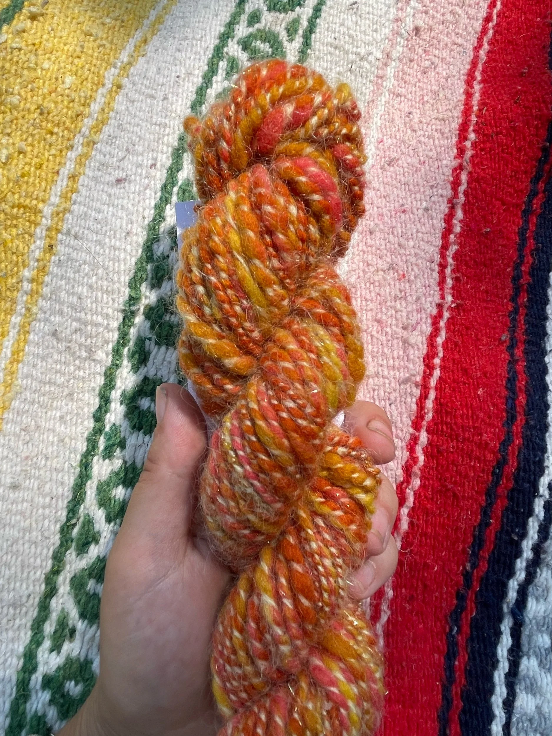 Hand holding a skein of multicolored, twisted yarn in orange, yellow, and red hues, lying on a striped textile with yellow, green, white, red, and blue colors.