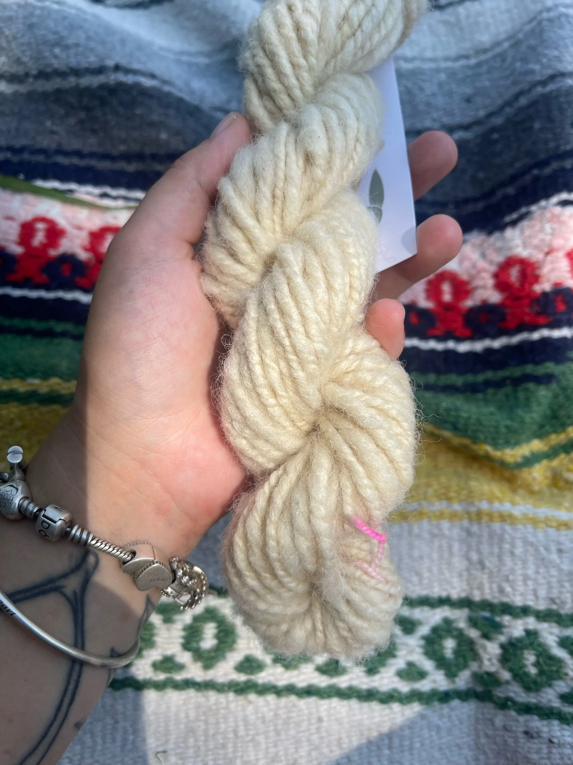 A hand holding a skein of thick, creamy white yarn, with a background of a colorful, patterned textile.