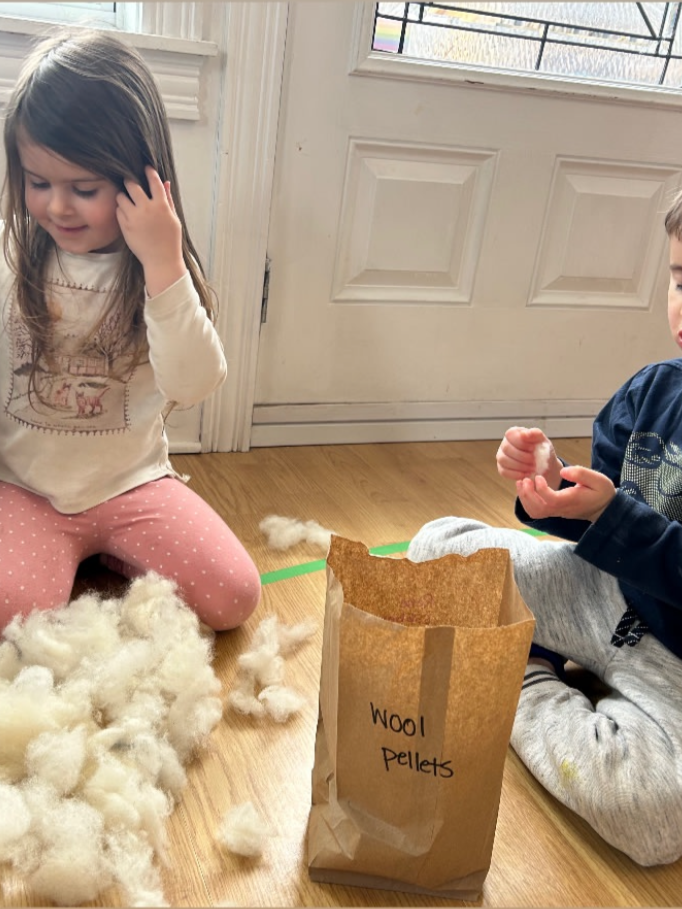 Two children sitting on a wood floor near a door, with a paper bag labeled "Wool Pellets" and piles of wool fibers around them, engaged in a sustainable sensory craft activity.