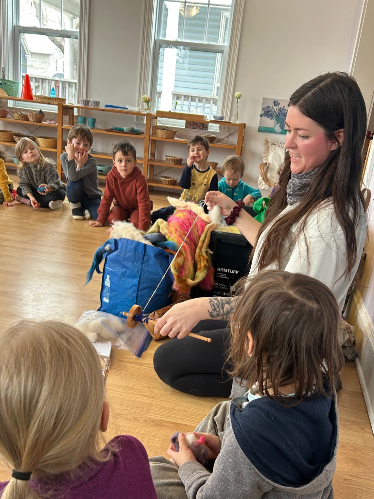 A woman is teaching children about fiber arts and spinning wool with colorful wool in a classroom. The children are sitting on the wooden floor, watching attentively.