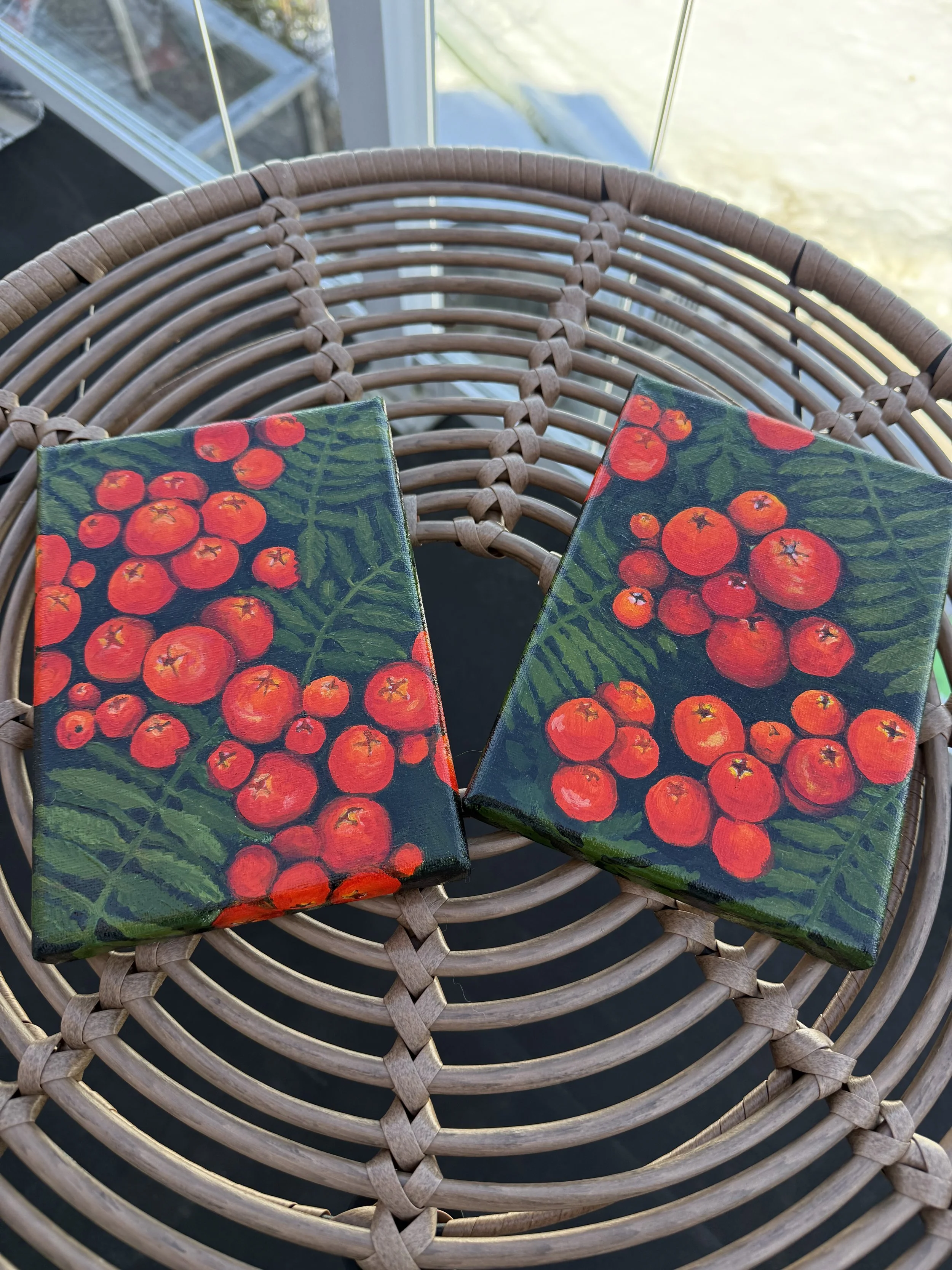 Two canvas prints with bright orange-red rowan berries and green leaves on a woven wicker chair, near a window with sunlight.