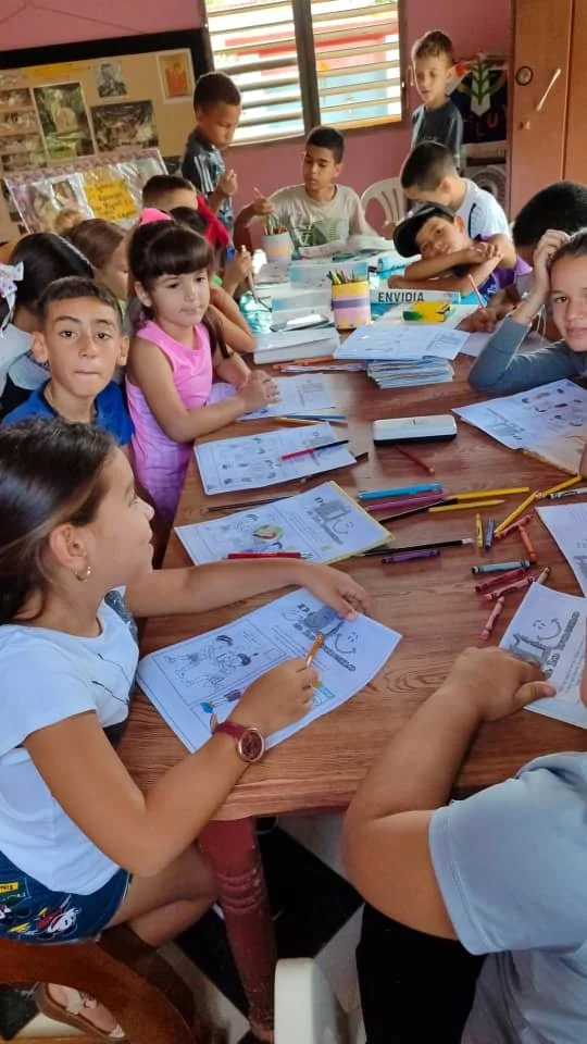 A classroom full of young children sitting around a table working on coloring pages with crayons and colored pencils, some children are looking at the camera while others focus on their work.