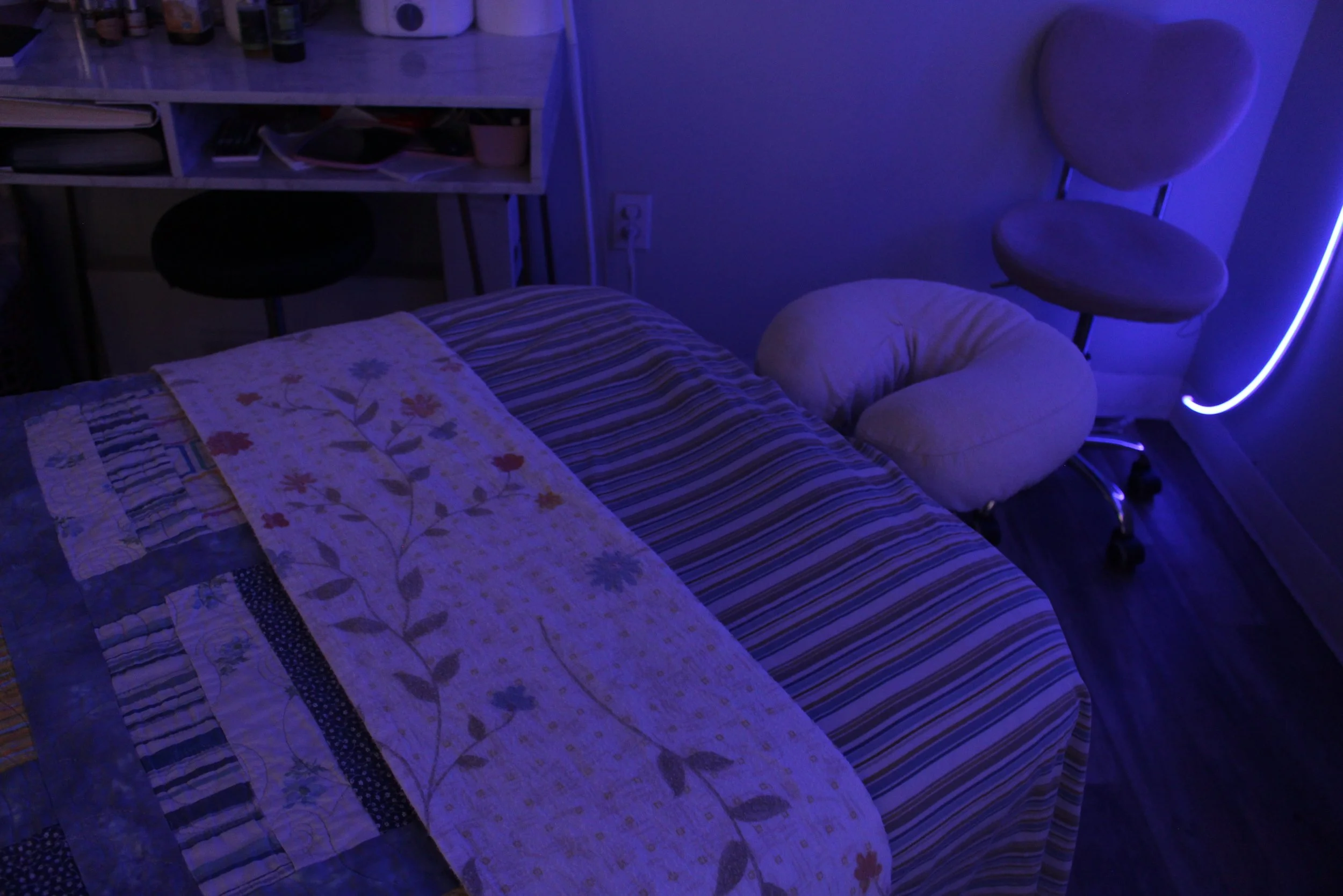 A treatment room with a massage table covered in a floral quilt, a striped blanket, a beige face cradle, a plush chair, a small desk with books and containers, and a blue LED strip light illuminating the room.