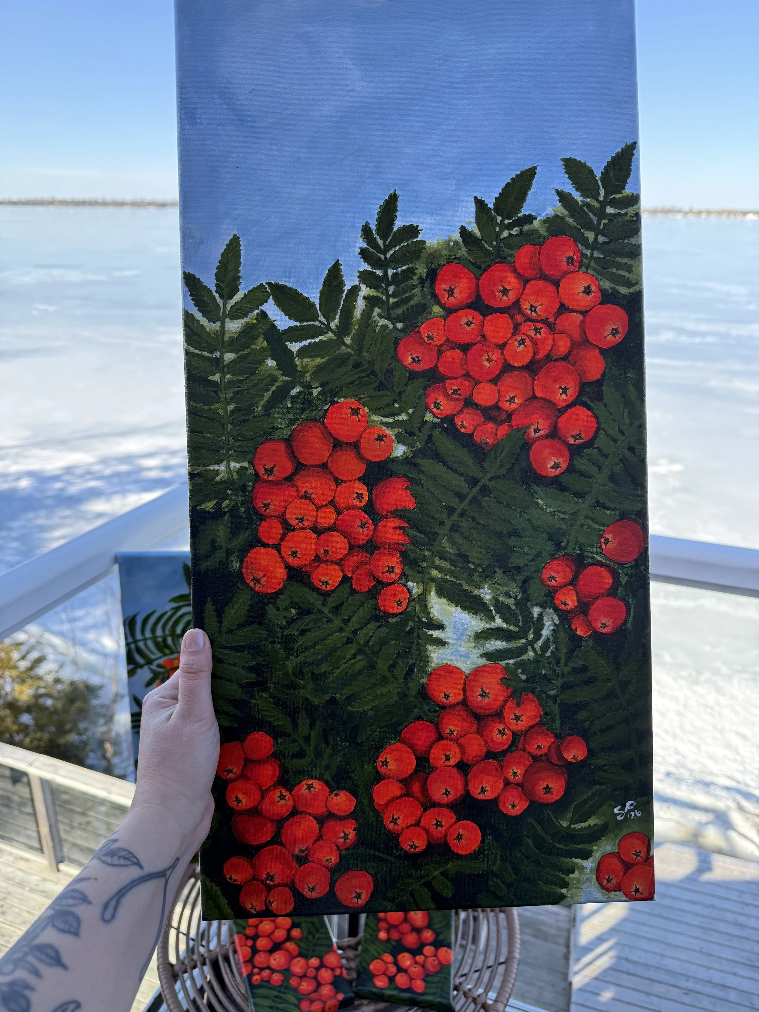 Hand holding a vertical painting of a cluster of red rowan berries with green leaves on a bright outdoor deck with a glass enclosure and a snowy landscape in the background.