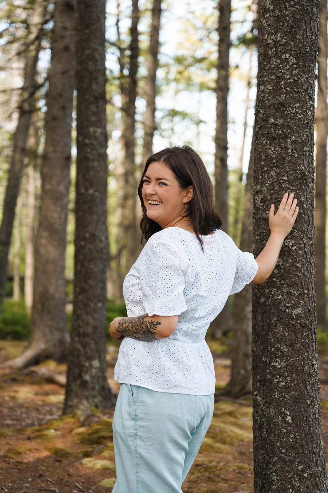 A woman with dark hair, smiling, leaning against a tree in a forest. She is wearing a white eyelet top and light blue pants, with a visible tattoo on her forearm.