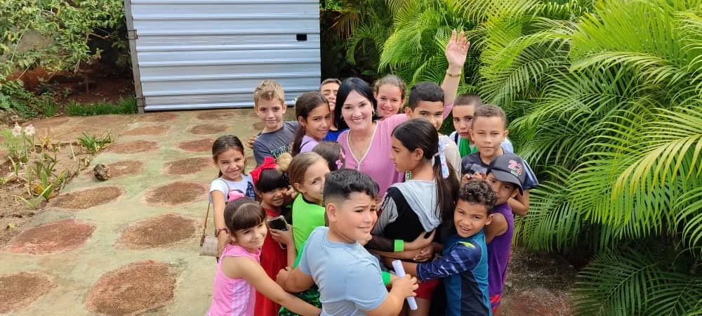 A group of children and a woman are gathered outdoors, smiling and making a circle around the woman, who is waving. The children are of various ages and are standing on a stone pathway with green plants and trees in the background.
