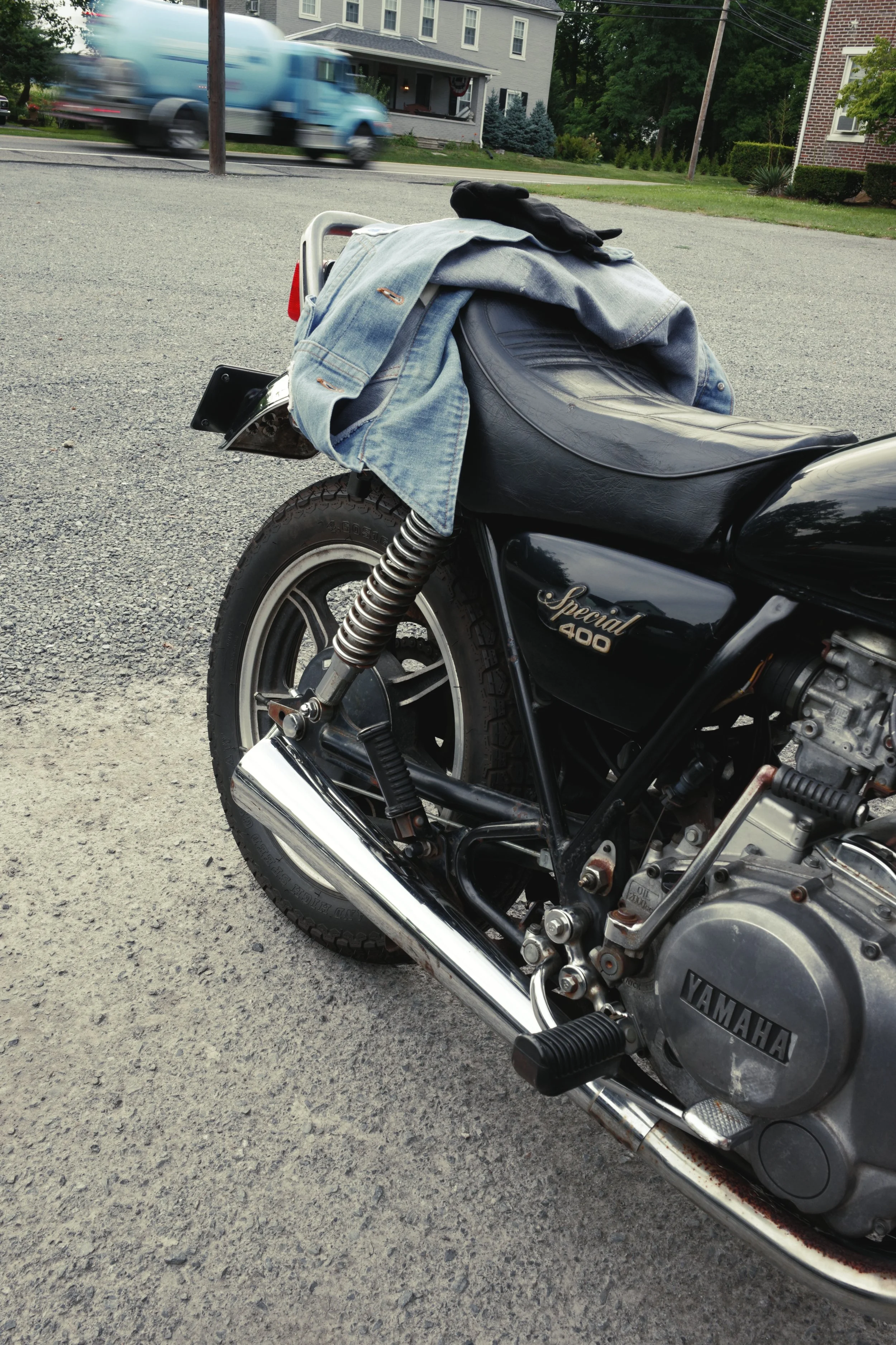 Close-up of a black Yamaha Special 400 motorcycle parked on gravel, with a denim jacket, black glove, and helmet placed on the seat. In the background, a blue truck is passing by on a street in a residential neighborhood with houses and trees.