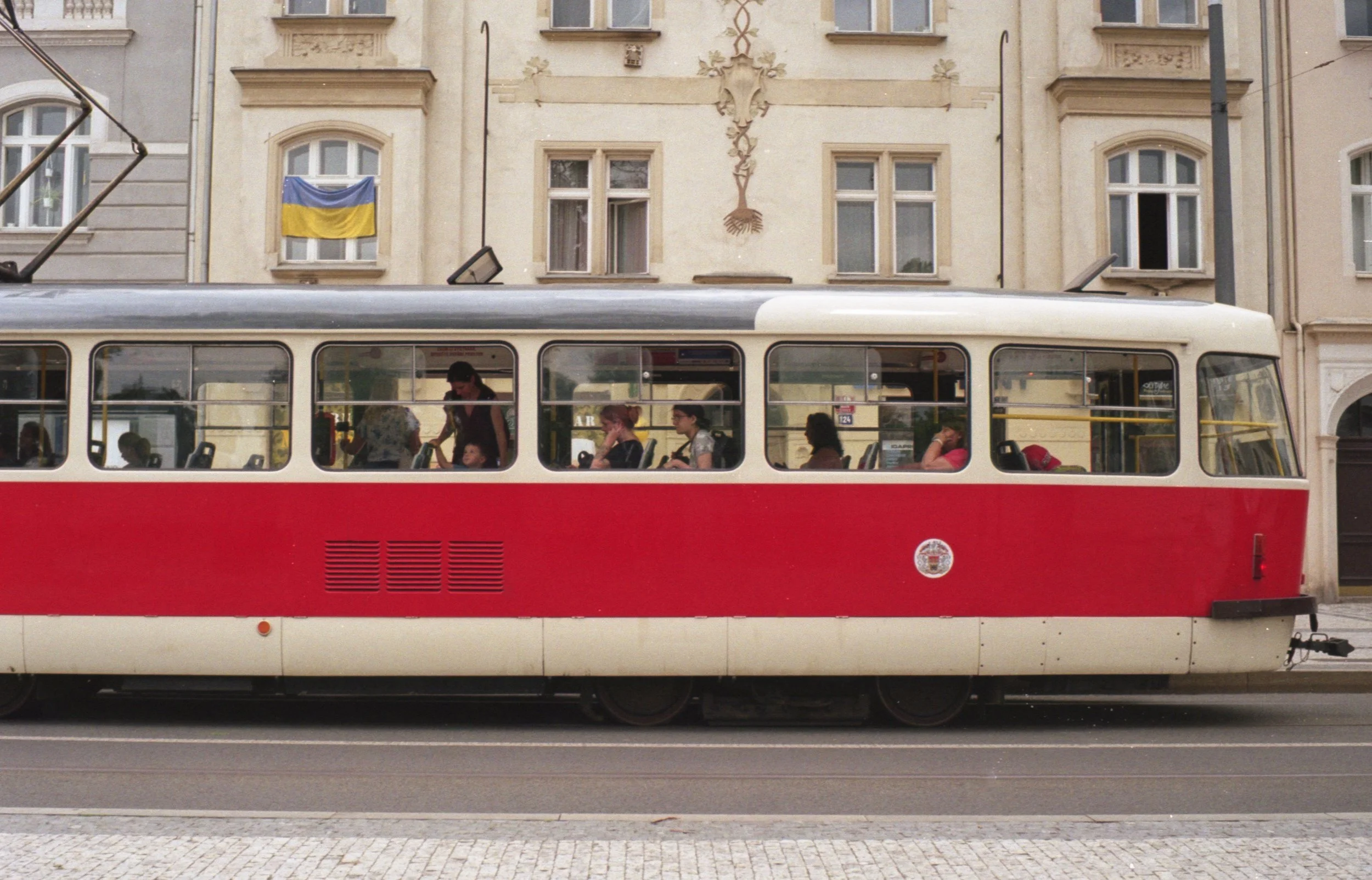 A red and white tram moving along a street with a building in the background. The building has a Ukrainian flag hanging from one window.