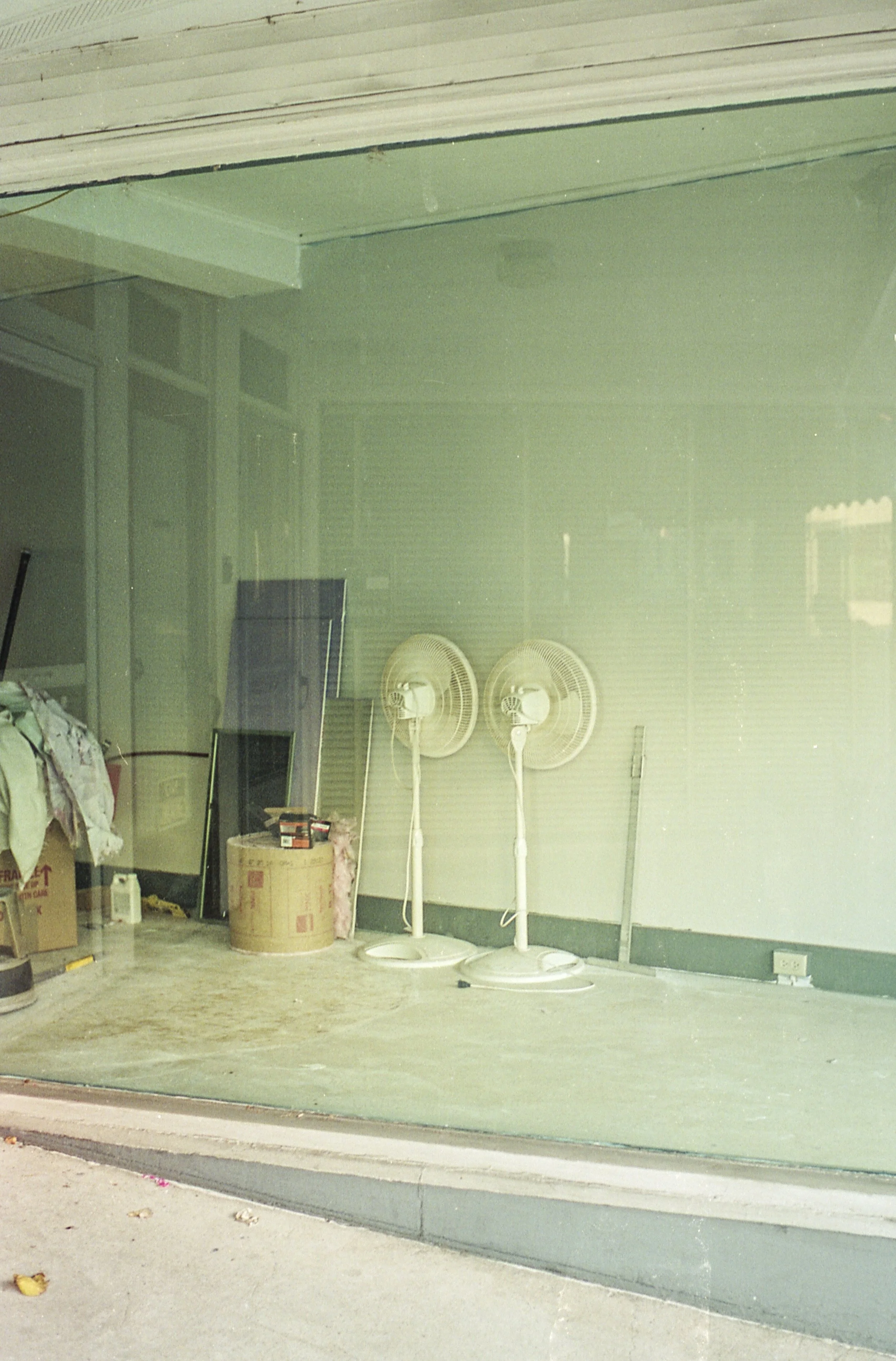 A storefront window displays two white oscillating fans, a large cardboard box, a small black framed television, some boxes, a white bottle, and a plastic bag inside. The window reflects some trees and the street outside, with a sidewalk and a curb v