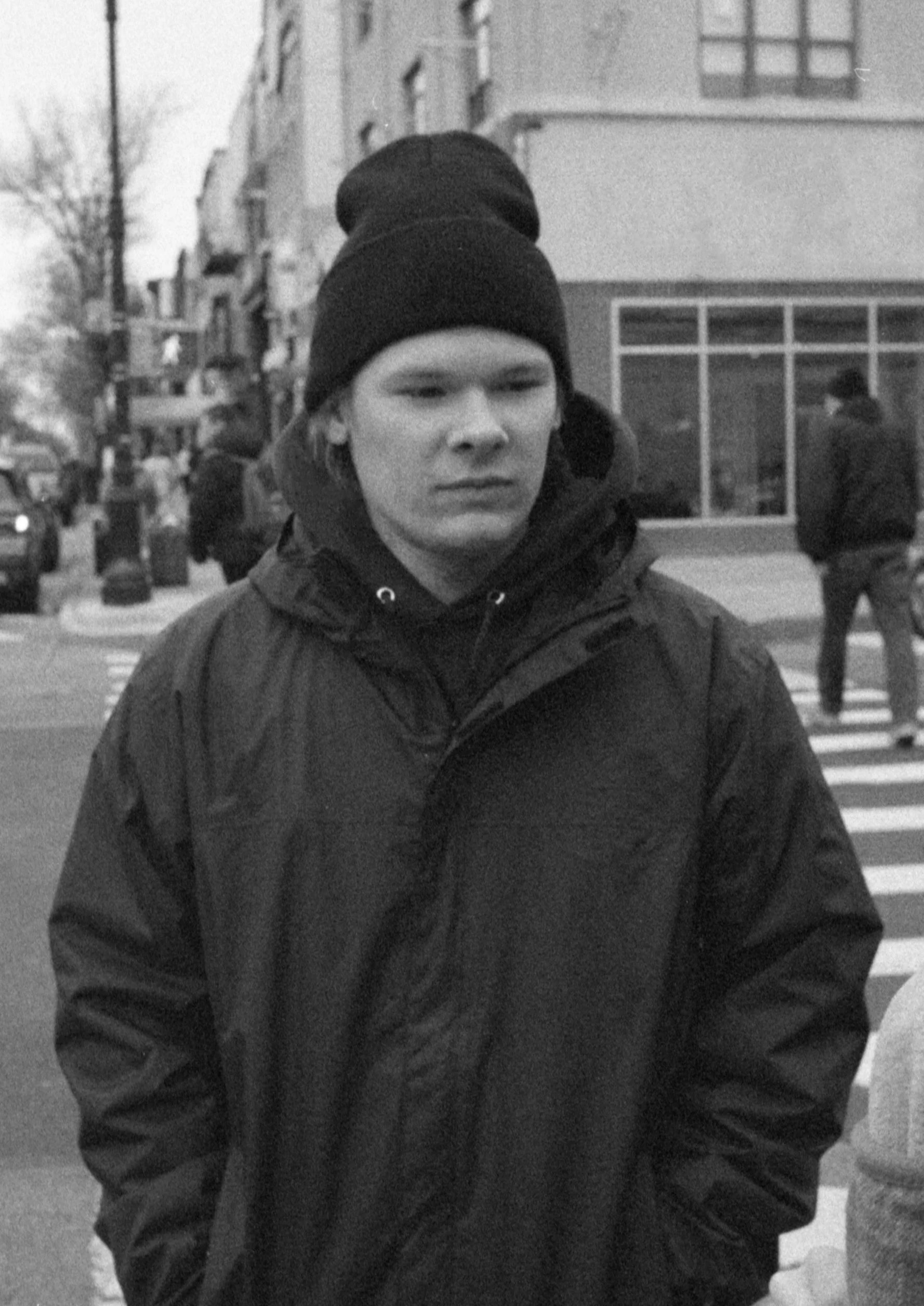 A person wearing a knit hat and a thick winter jacket standing on a city street with buildings and other pedestrians in the background.