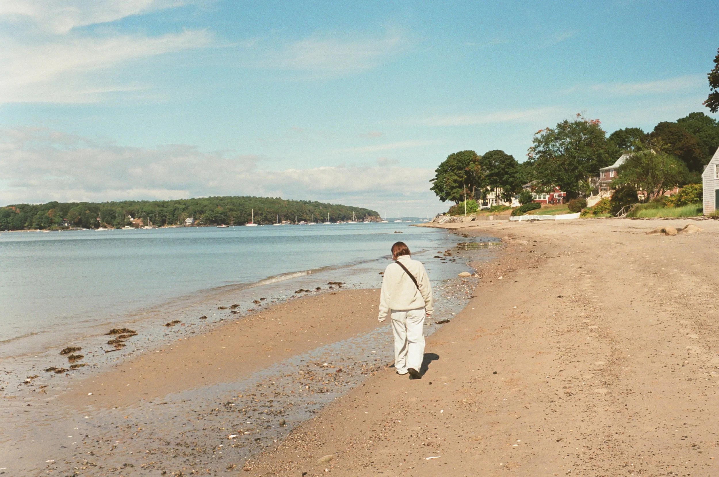 A person wearing a white hooded jacket and light-colored pants walking along a sandy beach near the water with houses and trees in the background under a partly cloudy sky.