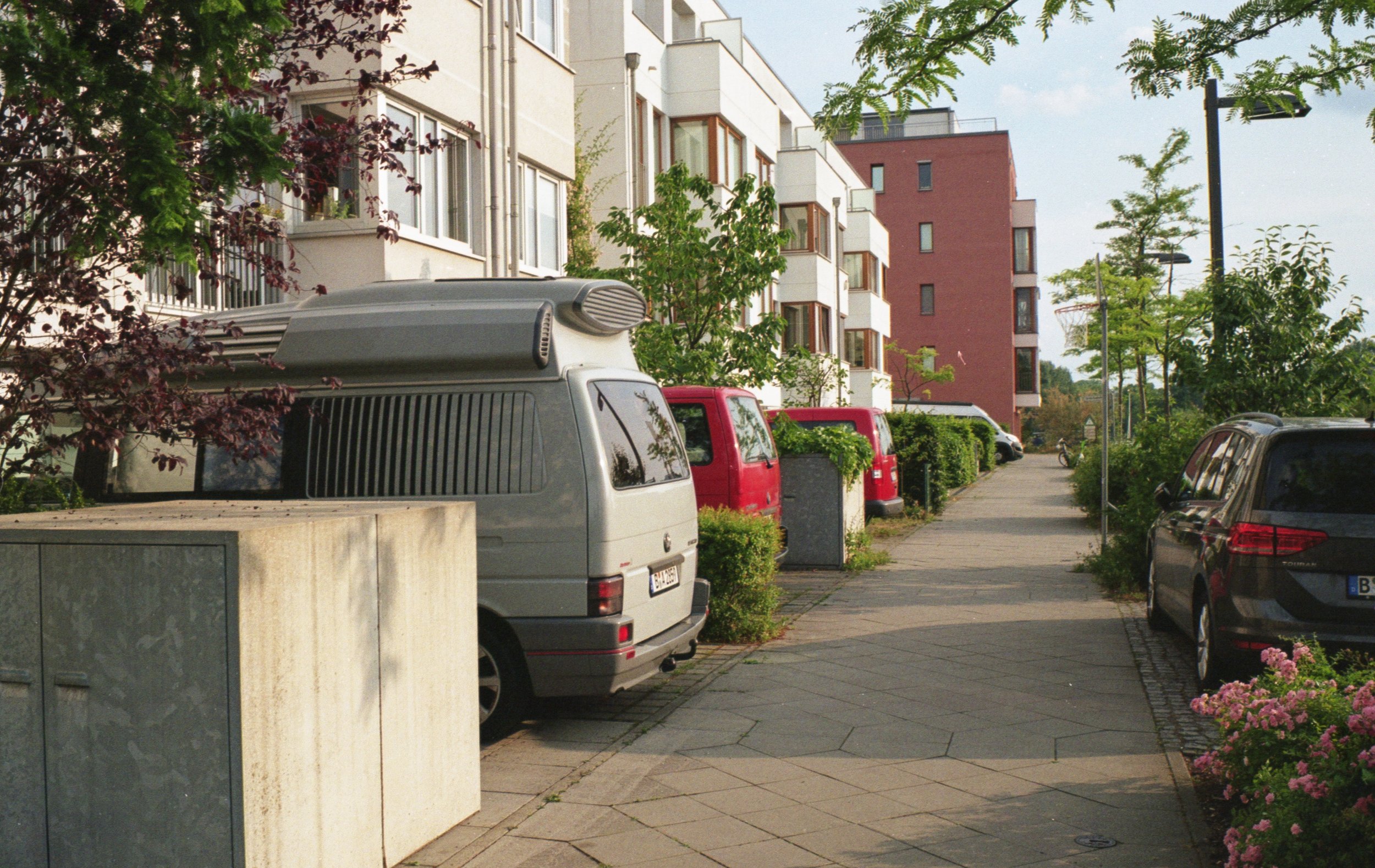 A residential street with parked cars, modern apartment buildings, trees, and flowering plants.