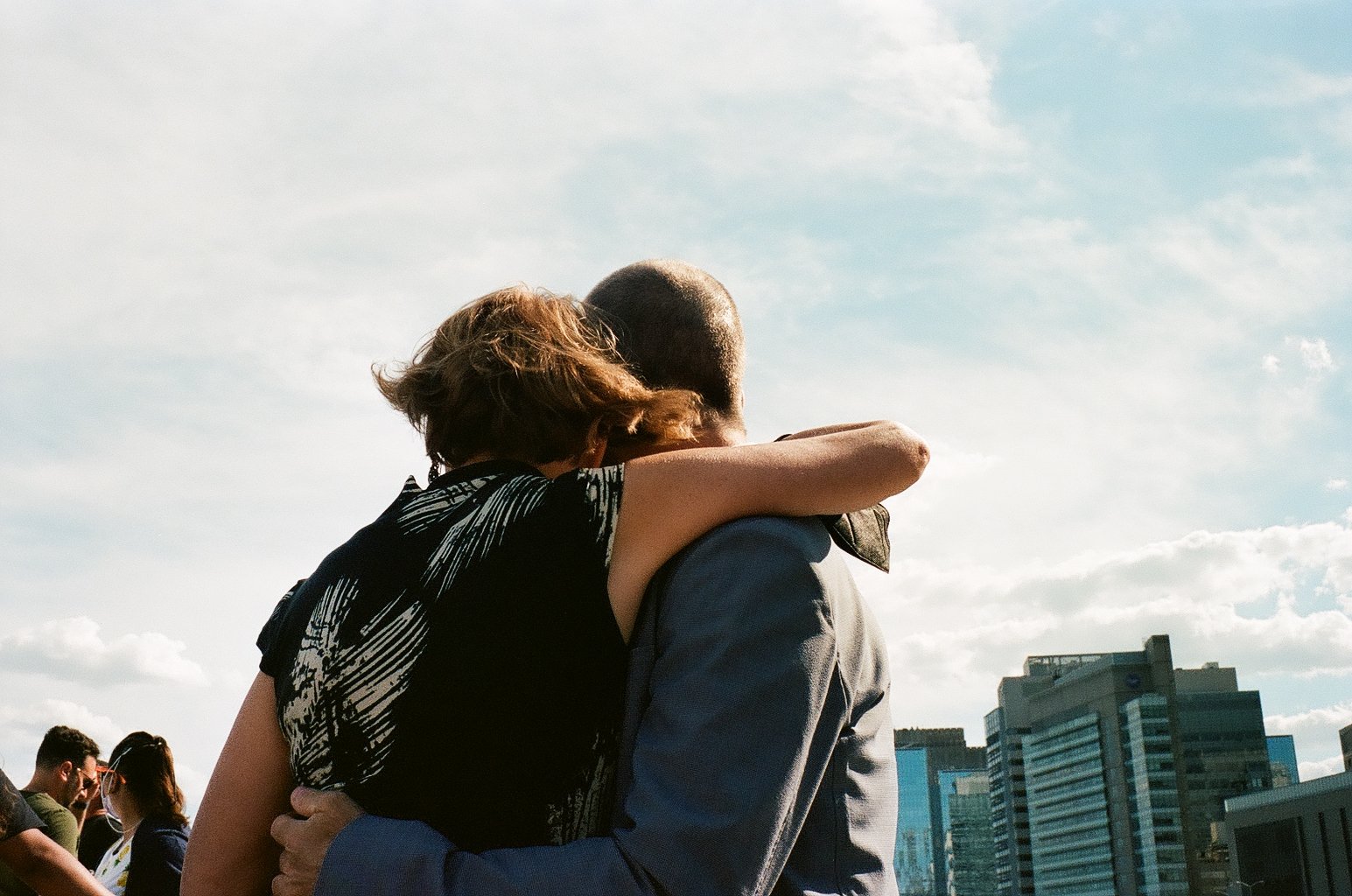 A woman with short hair and a man are embracing outdoors, with the woman’s arm around the man’s neck. They are in an urban setting with buildings and other people in the background, under a partly cloudy sky.