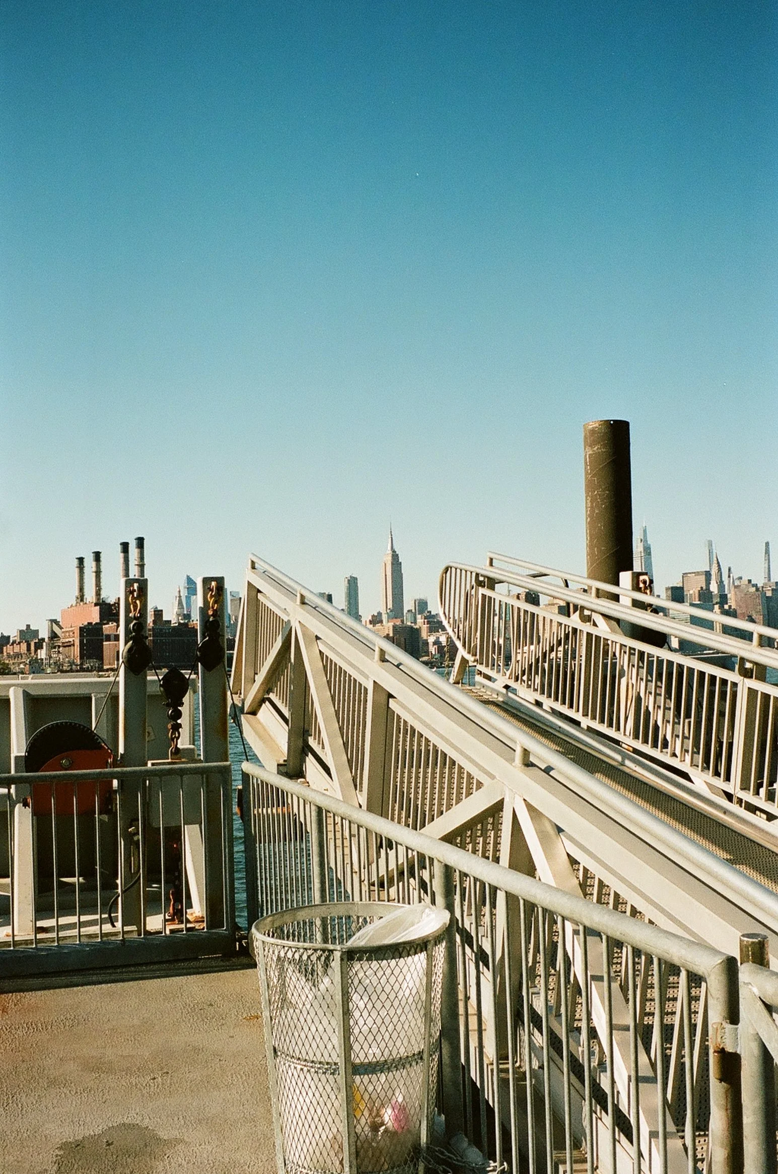 View of a city skyline, including the Empire State Building, from a ferry deck with metal railings, a trash can, and a structure of stairs and safety barriers in the foreground, under a clear blue sky.