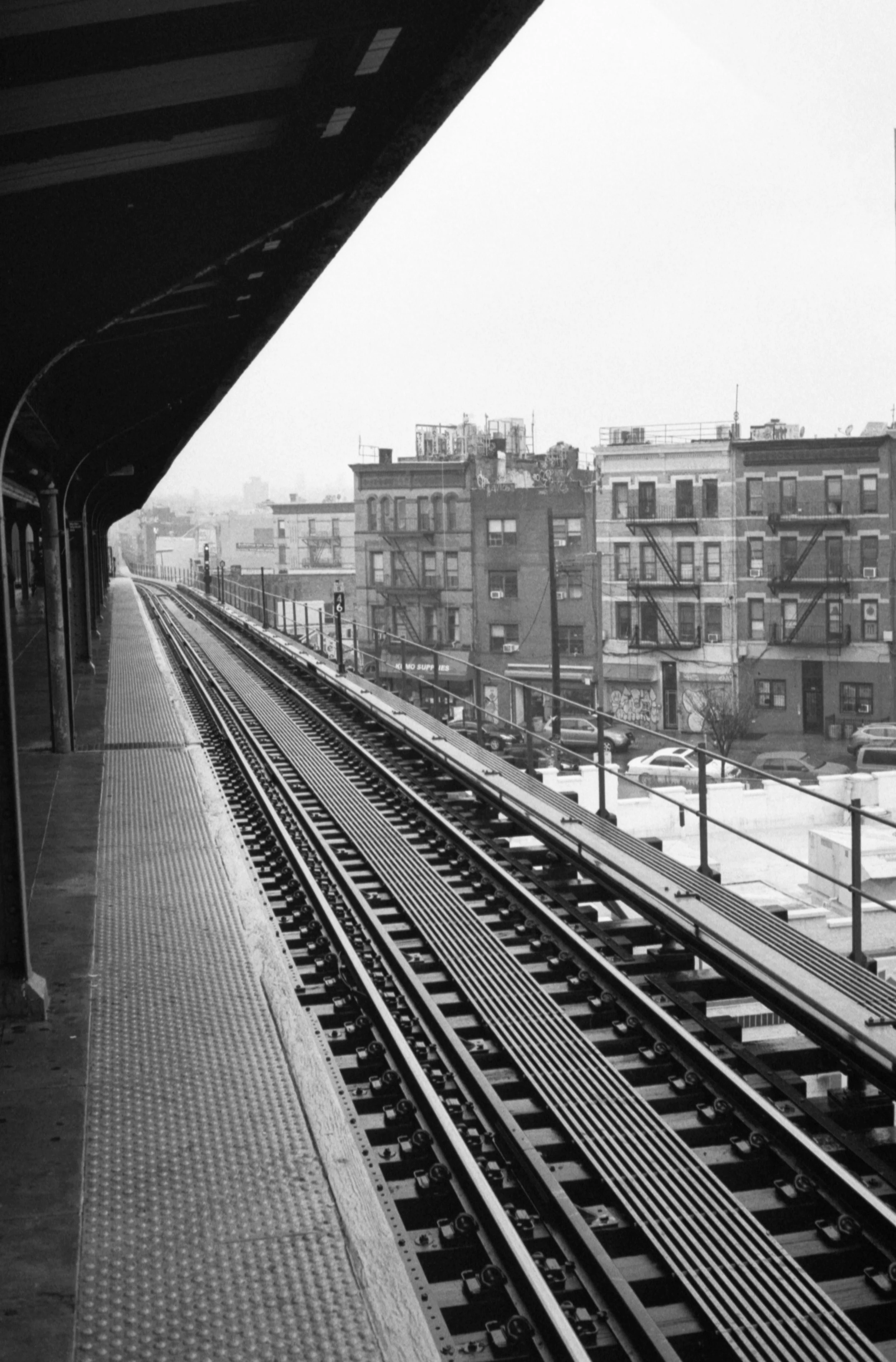 View of an elevated train station platform with tracks and city buildings in black and white.