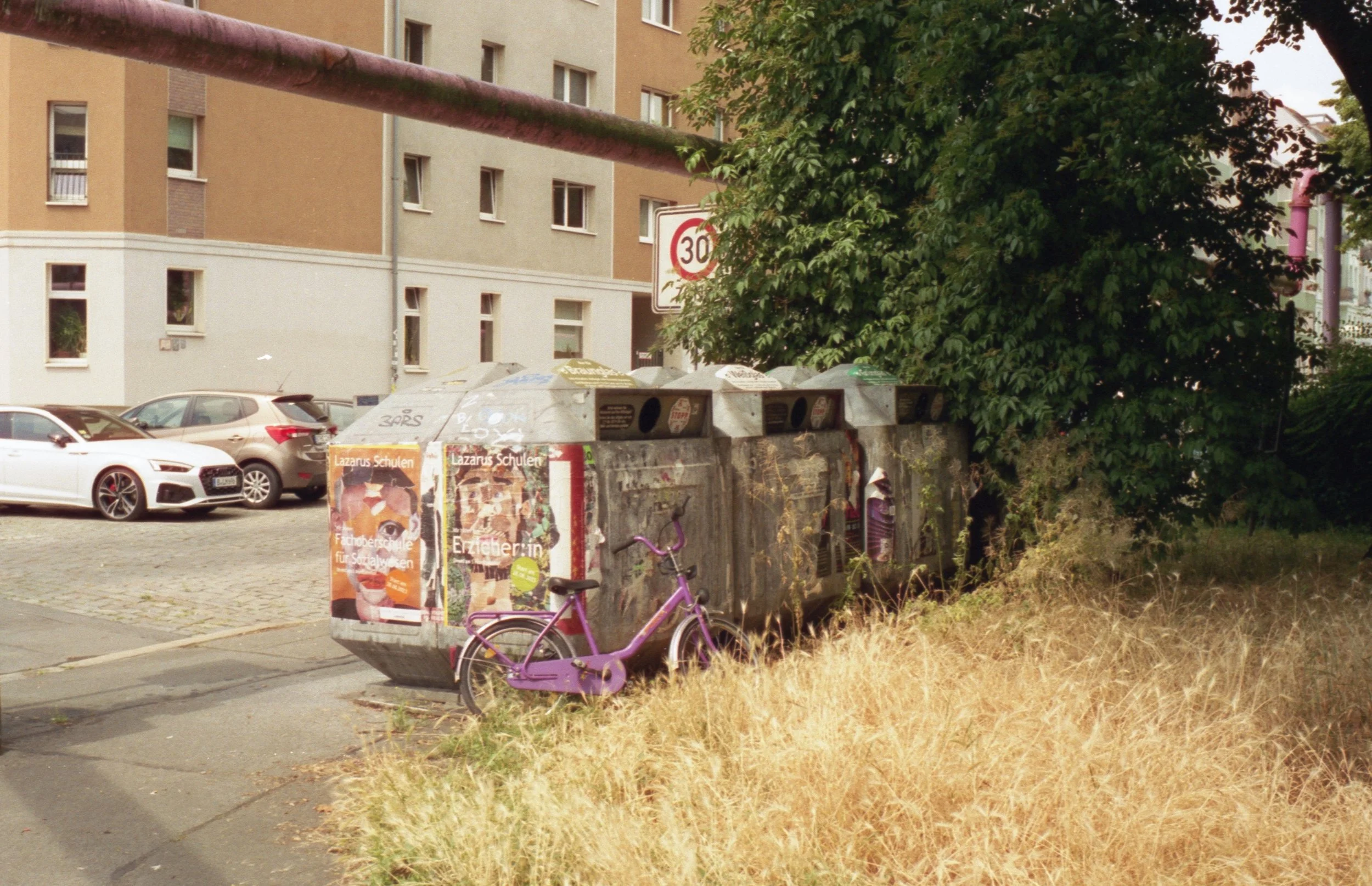 A purple bicycle leaning against a trash bin on a sidewalk. The trash bin has advertisements and graffiti on it. Behind it are parked cars and an apartment building. A large green tree partially covers the right side of the scene. There is a 30 km/h 
