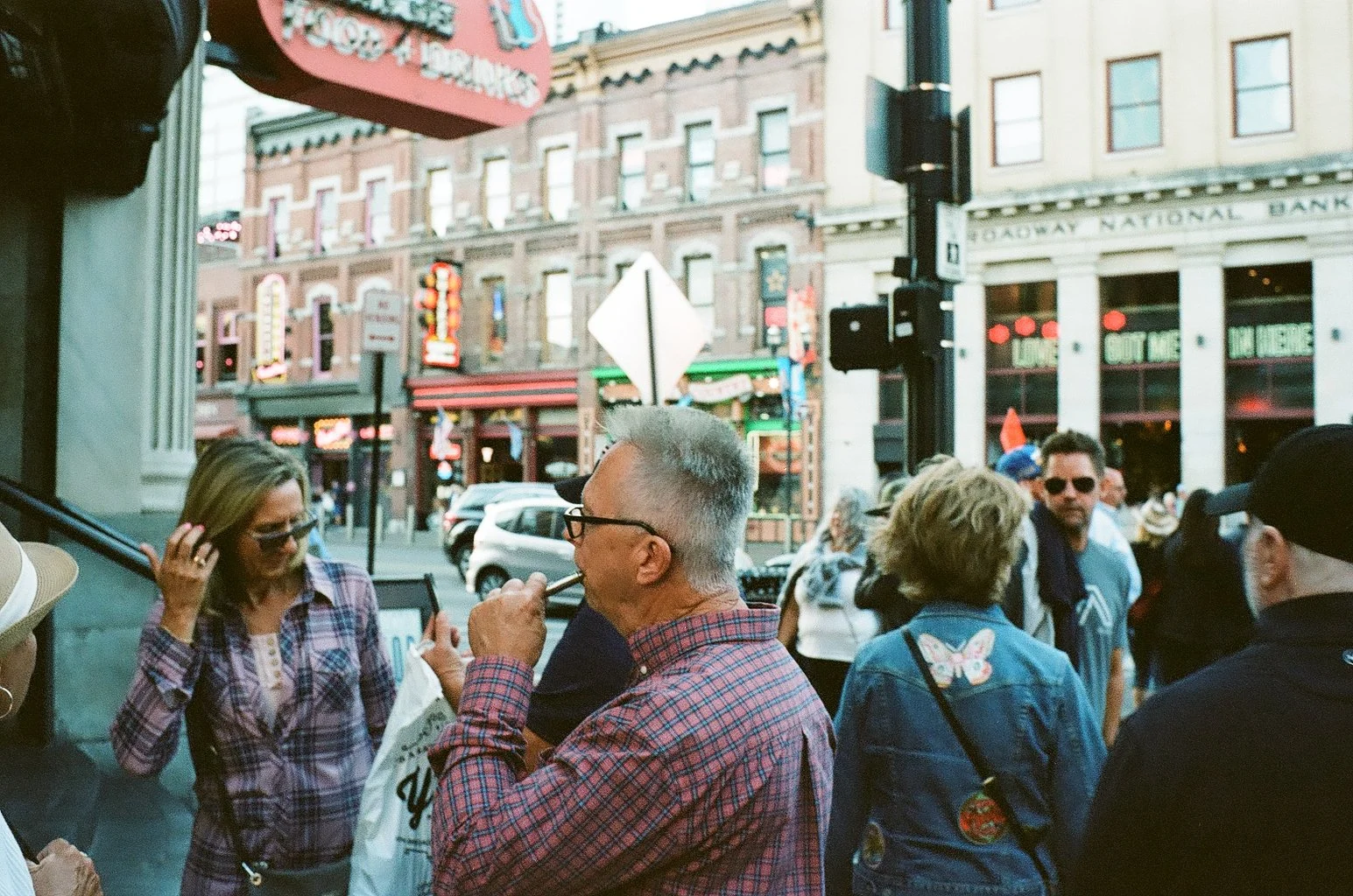 A group of people standing outside on a city street, engaging in conversation and taking photos, with historic buildings and neon signs in the background.