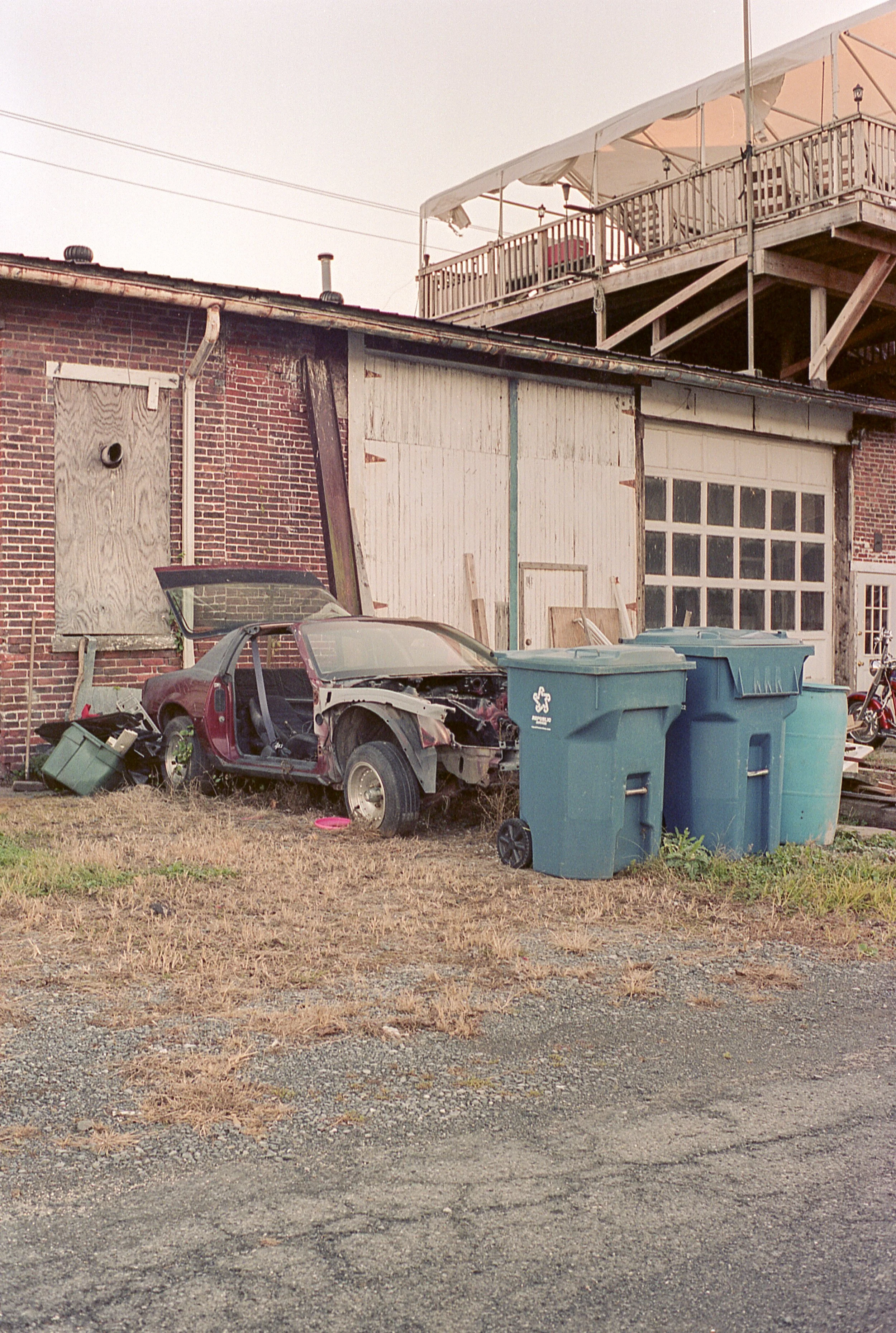 An old, damaged car with a missing door and front bumper is parked in front of a building with a brick wall, boarded-up window, and a white garage door. There are three large blue trash bins in the foreground. The building has a wooden second-story d