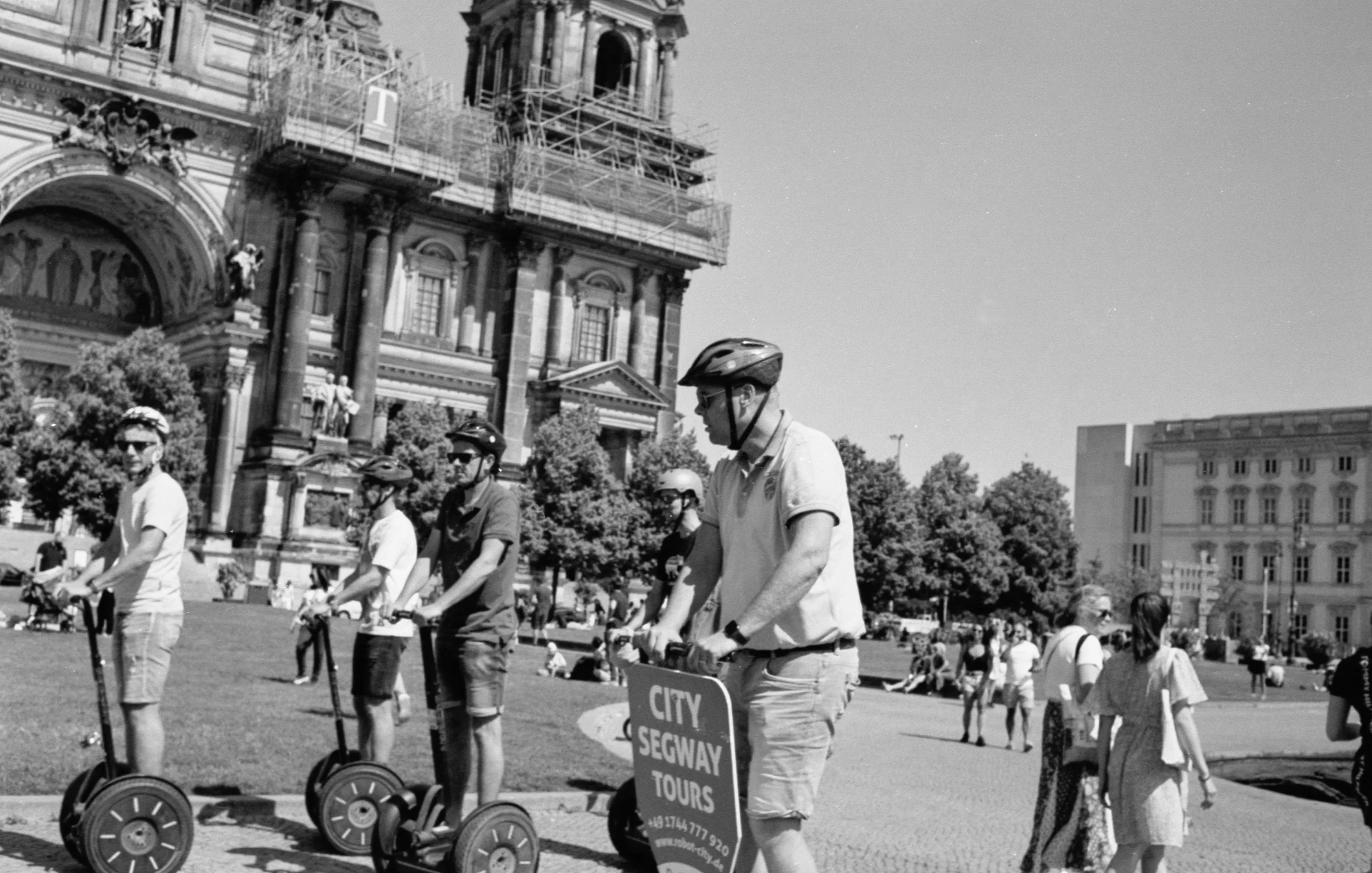 A group of people riding electric scooters in an urban park with historic buildings and a church tower in the background. People are wearing helmets and sunglasses, and there is a sign advertising city Segway tours.