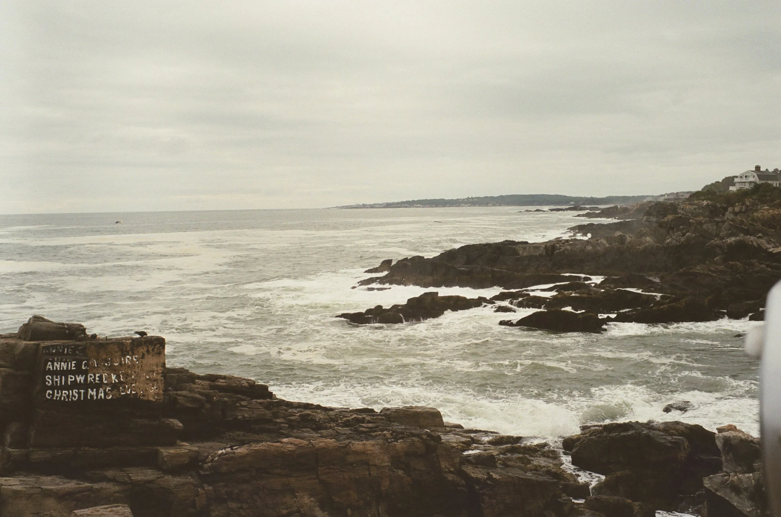 Overcast sky over rocky coastline with waves crashing against rocks and a house situated on a hill in the distance, with a sign in the foreground that reads, 'ANNE C. GARDEN SHIPWRECK CHRISTMAS'.